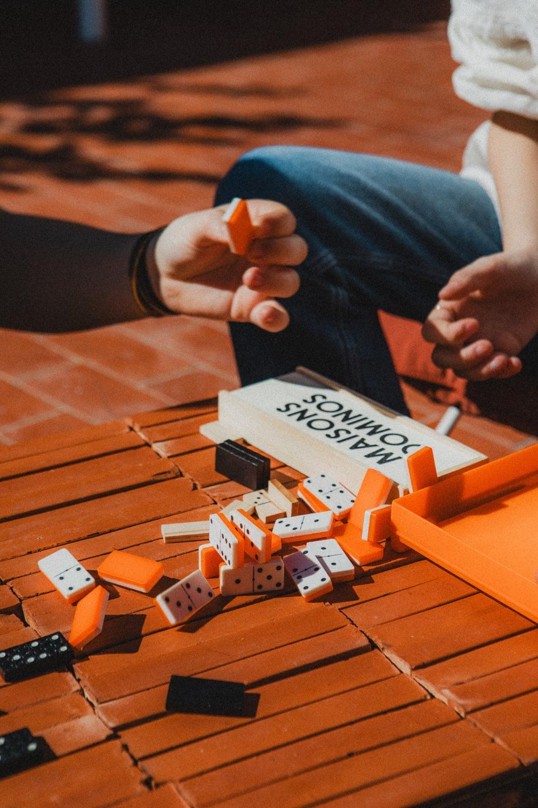 Two people are playing Dominoes at a wooden table. One person is holding a domino tile, and several tiles are scattered on the table. An orange container and a white booklet are next to them. The scene takes place in a bright outdoor space.