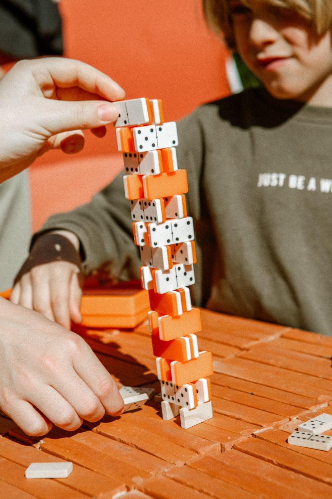 Two children are playing with orange and white Domino Deux, carefully stacking them to form a tall, precarious tower on a textured orange surface. One child's hand is stabilizing the tower while the other child looks on with a focused expression.