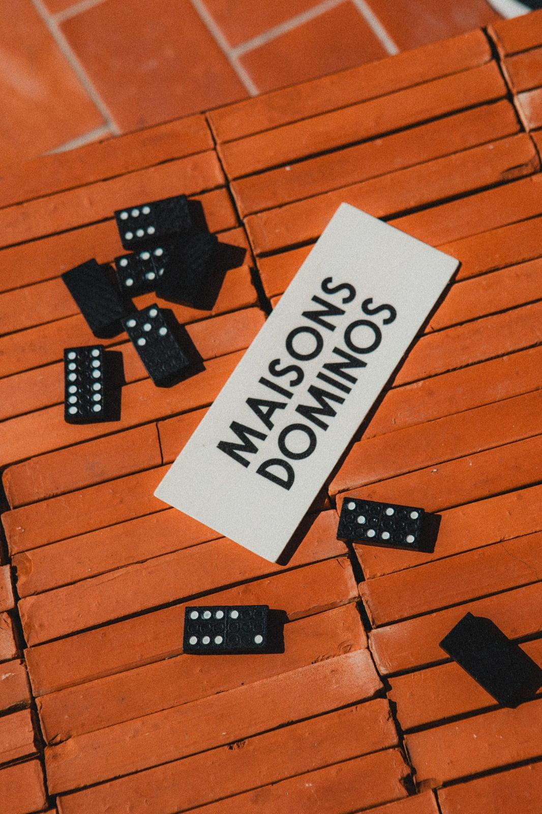 Un groupe de dominos noirs est dispersé sur une surface en brique rouge, entourant une carte rectangulaire blanche avec le texte « Domino Deux Maisons » en caractères gras noirs. L'image semble avoir été prise en plein jour.