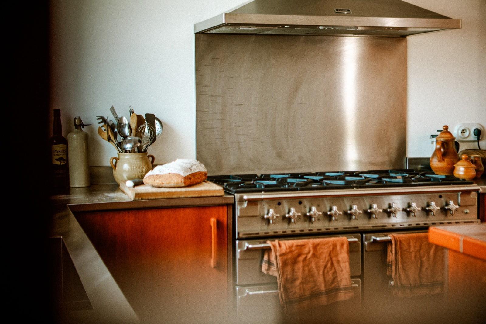 A cozy kitchen with a large stainless steel gas stove and oven dominating the space. Brown towels hang on the oven handle, echoing the elegant touches of Domino Deux. A loaf of bread rests on a cutting board next to a ceramic pitcher filled with utensils. The backsplash and stove hood are also stainless steel.