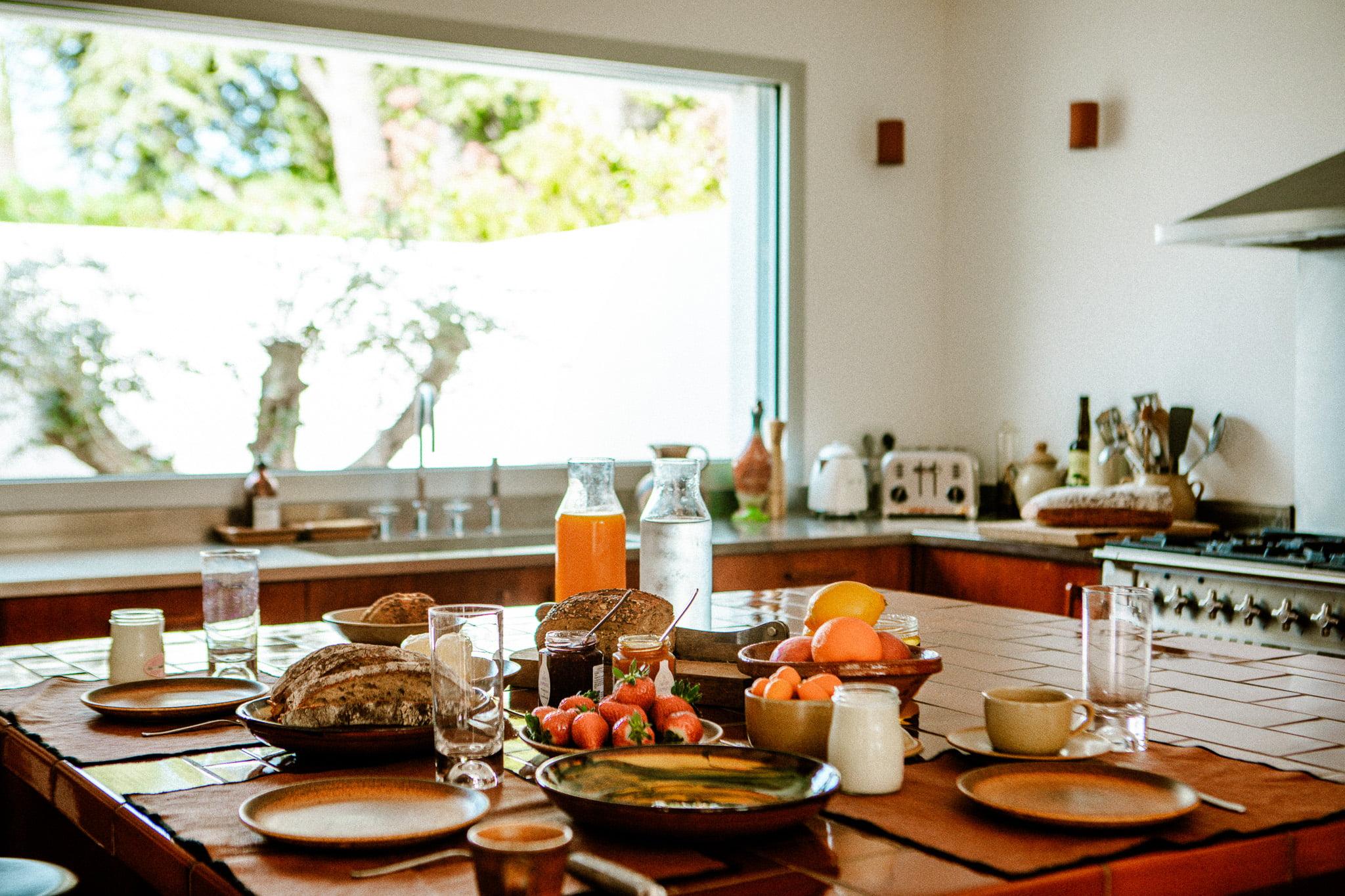 A kitchen table is set with a variety of foods, including fresh bread, strawberries, oranges, and glass pitchers containing water and juice. The background features a large window overlooking greenery, with appliances and kitchen utensils neatly arranged on the countertops.