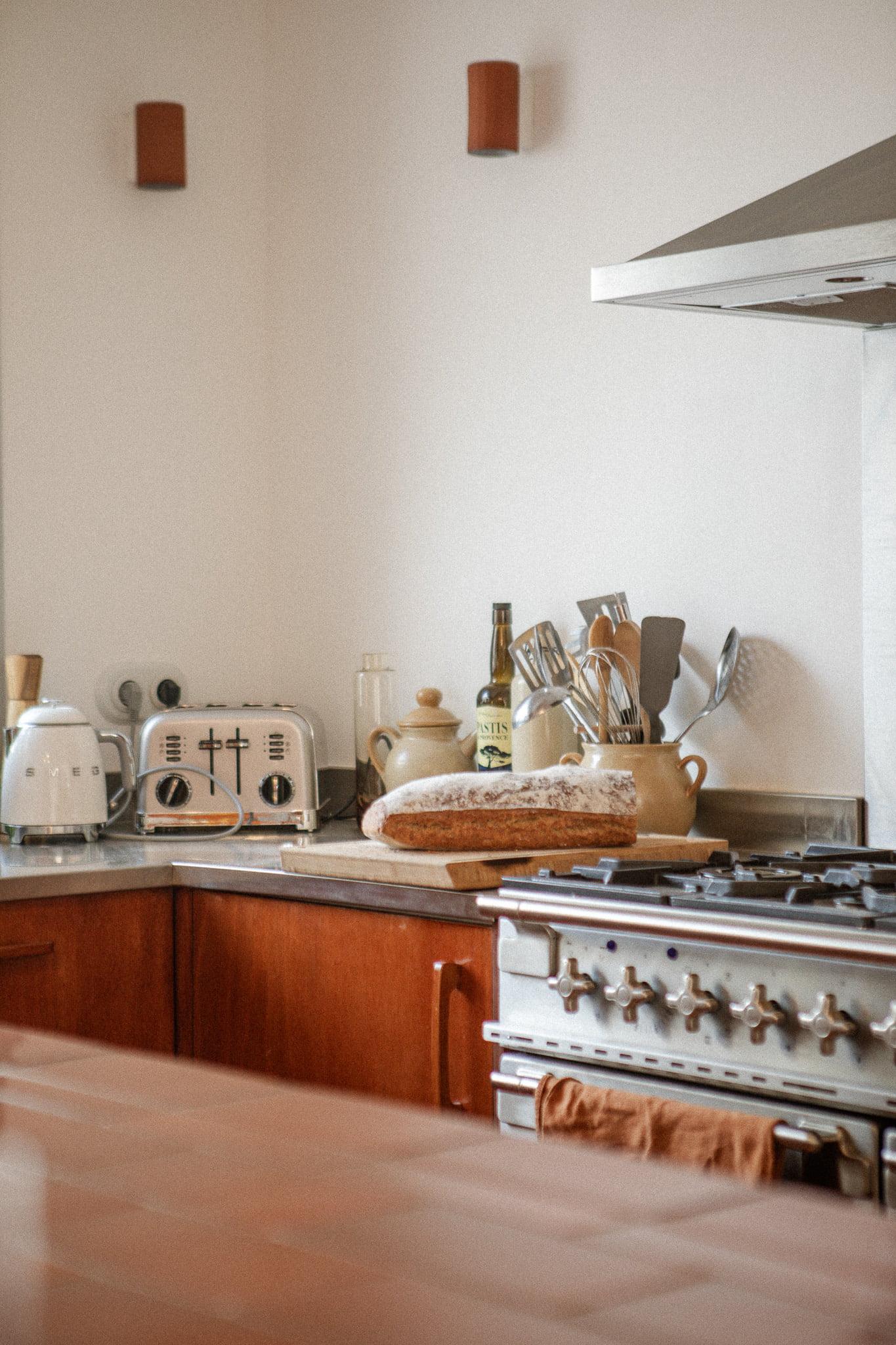 A cozy kitchen with wooden cabinets and stainless steel appliances. A baguette is resting on a wooden cutting board on the counter next to a toaster and various utensils. An orange towel hangs over the handle of the oven door on the stove. Warm lighting fixtures adorn the wall.