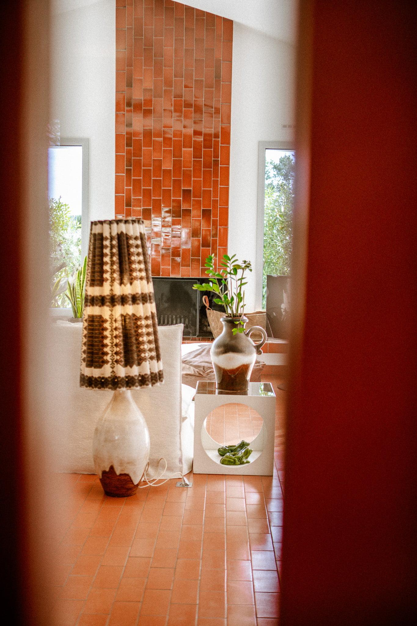 A warm and sunny living room with a patterned lampshade floor lamp, large ceramic vases, a white side table with a small plant, and a fireplace with red tiles. The room features a terracotta floor and large windows revealing the greenery outside.