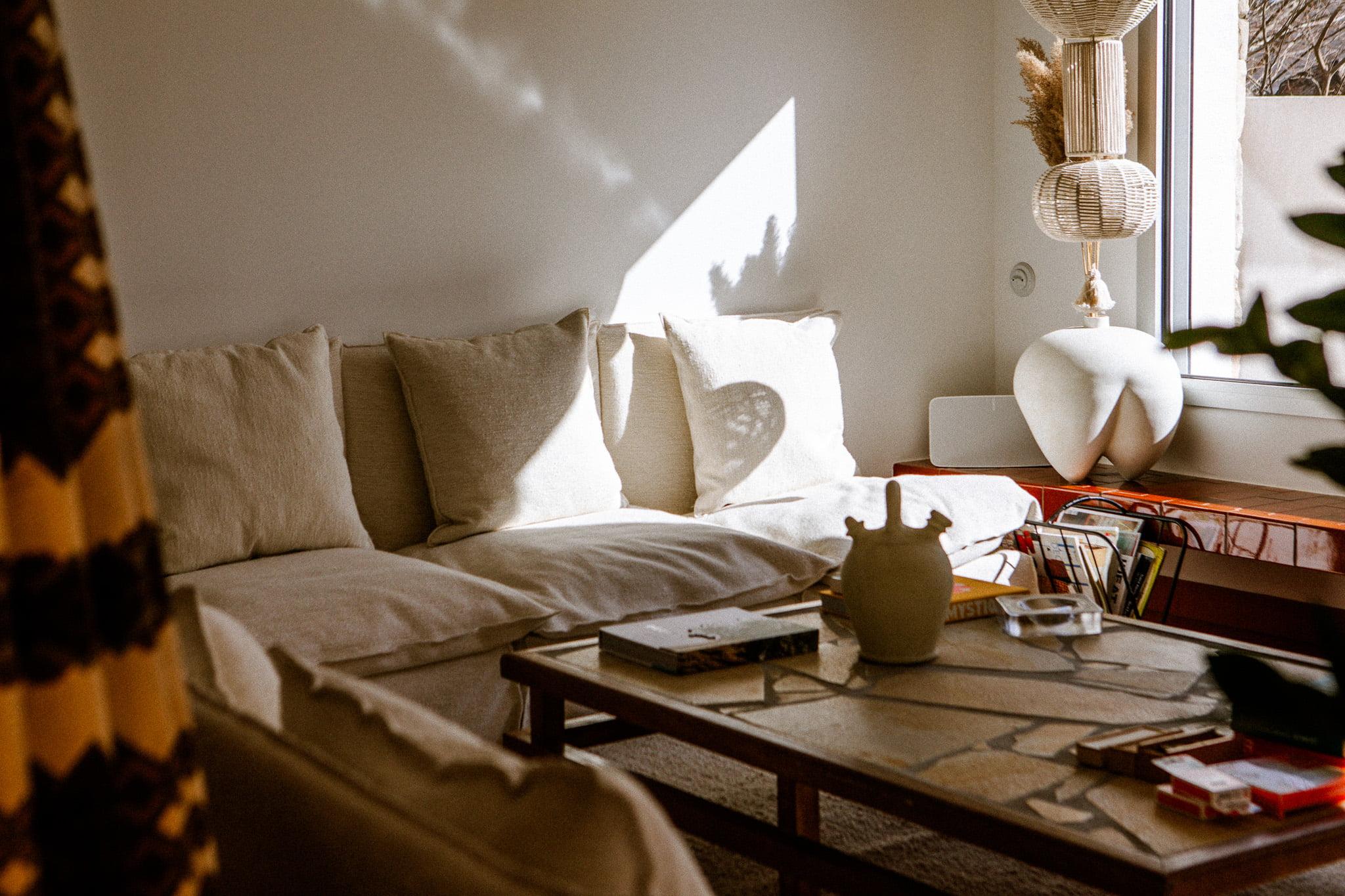 A warm living room bathed in natural light features a beige sofa with white cushions, a wooden coffee table with books and decorative items, and a large window letting in light. A sculpted white lamp and decorative objects are visible on a side table next to the sofa.