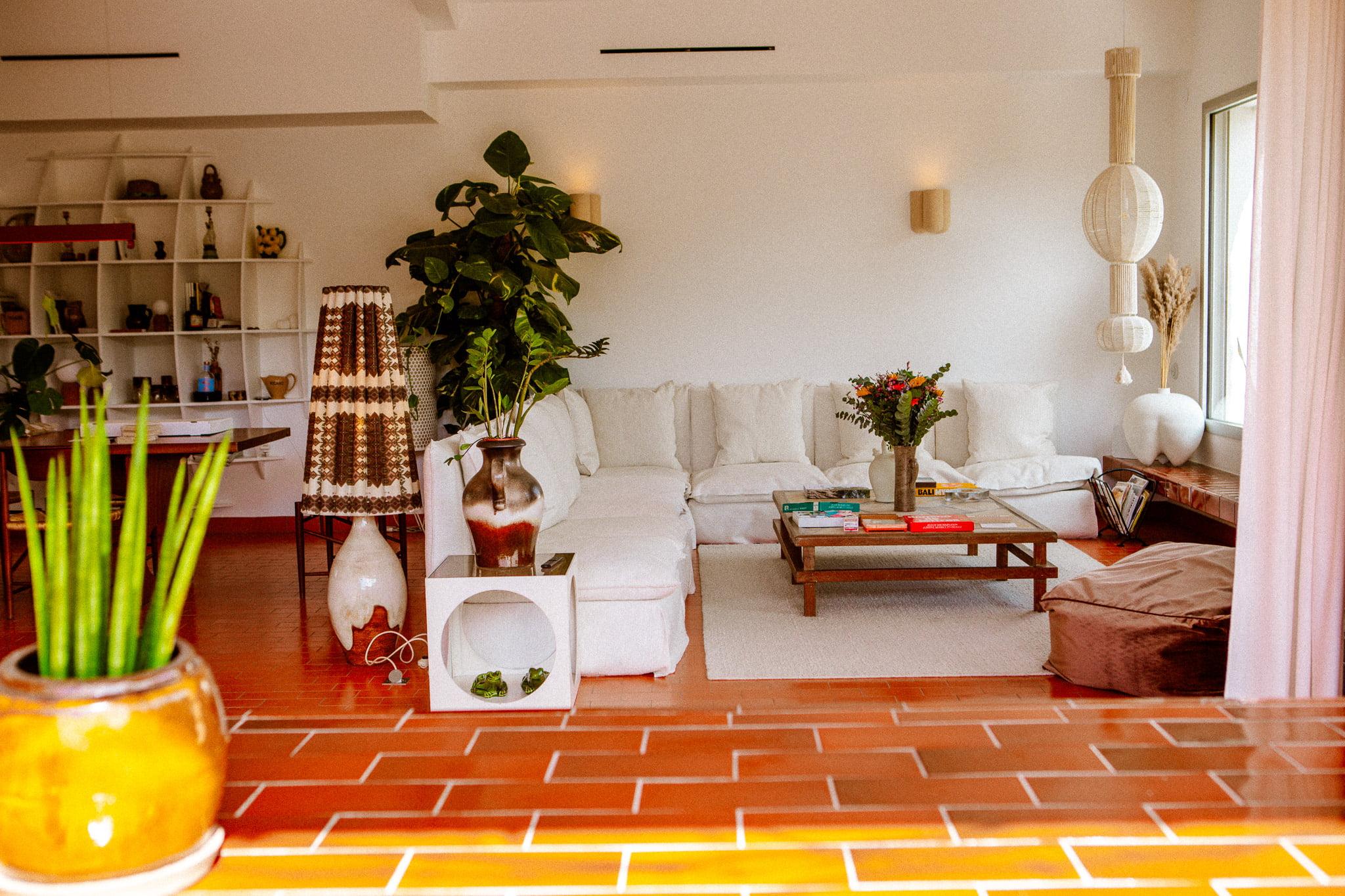 Un salon confortable et bien éclairé avec un grand canapé d'angle blanc orné de coussins colorés. Une table basse en bois avec un vase de fleurs se trouve devant. La pièce est agrémentée d'un éventail de plantes, d'une décoration moderne et d'une bibliothèque murale.