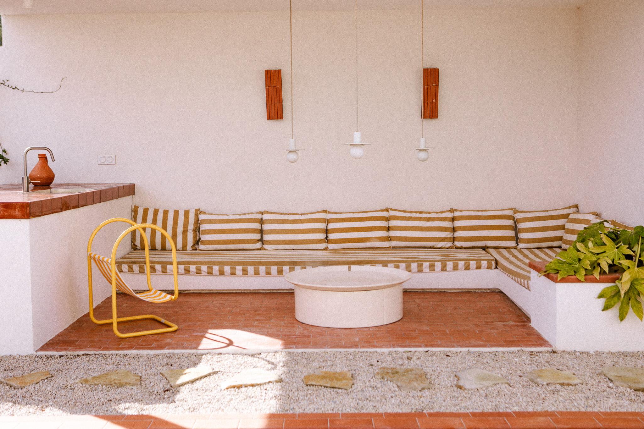 A cozy patio with a long striped cushioned bench, a round white coffee table, and a modern yellow chair. Potted plants decorate the corner, and three pendant lights hang above. The floor features a mix of red tiles and pebbles.