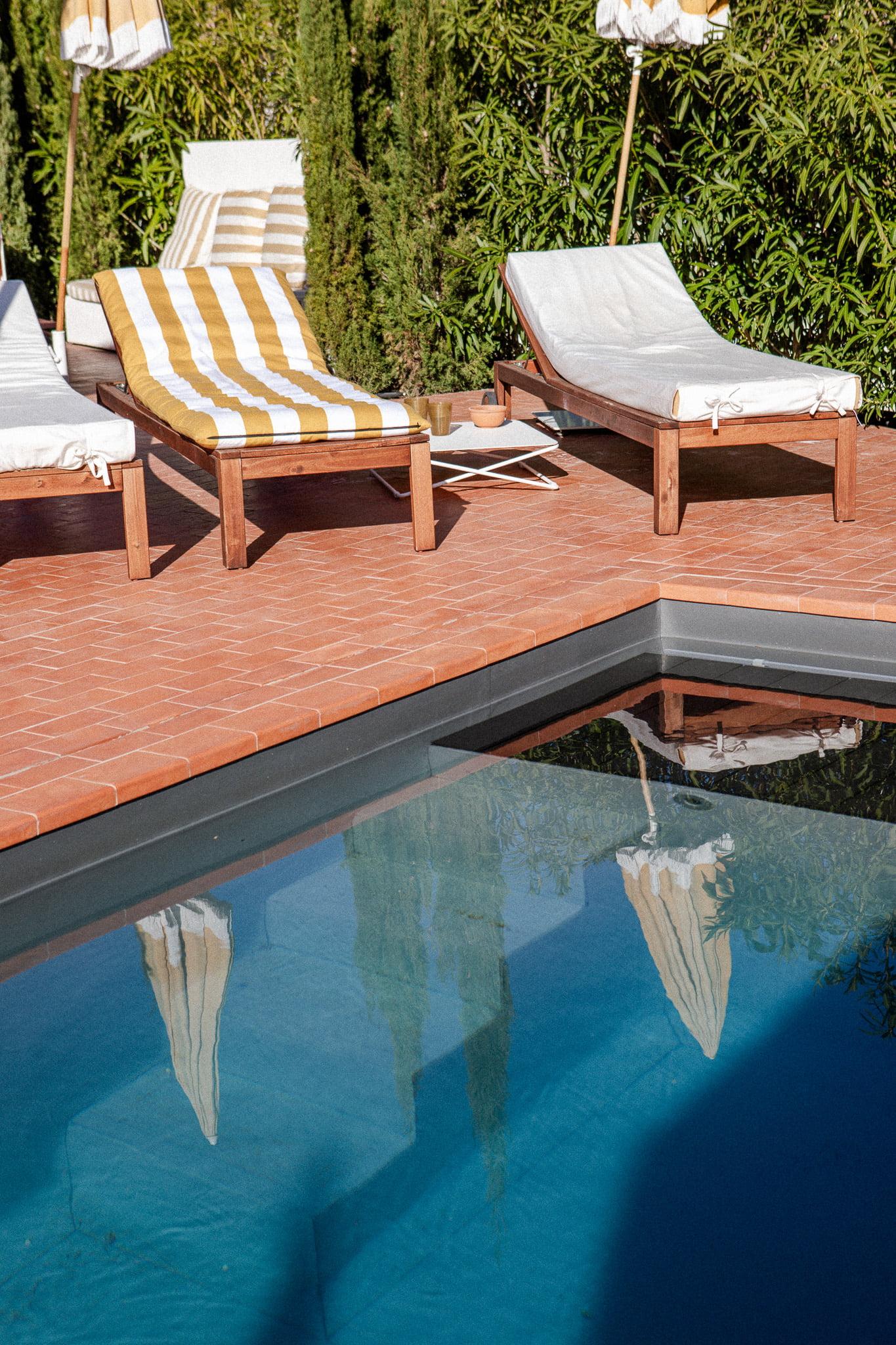 A serene poolside scene with several wooden lounge chairs featuring white and yellow striped cushions. The lounge chairs are arranged on a tiled deck with green shrubs in the background. The light blue pool reflects a sunny and peaceful atmosphere.