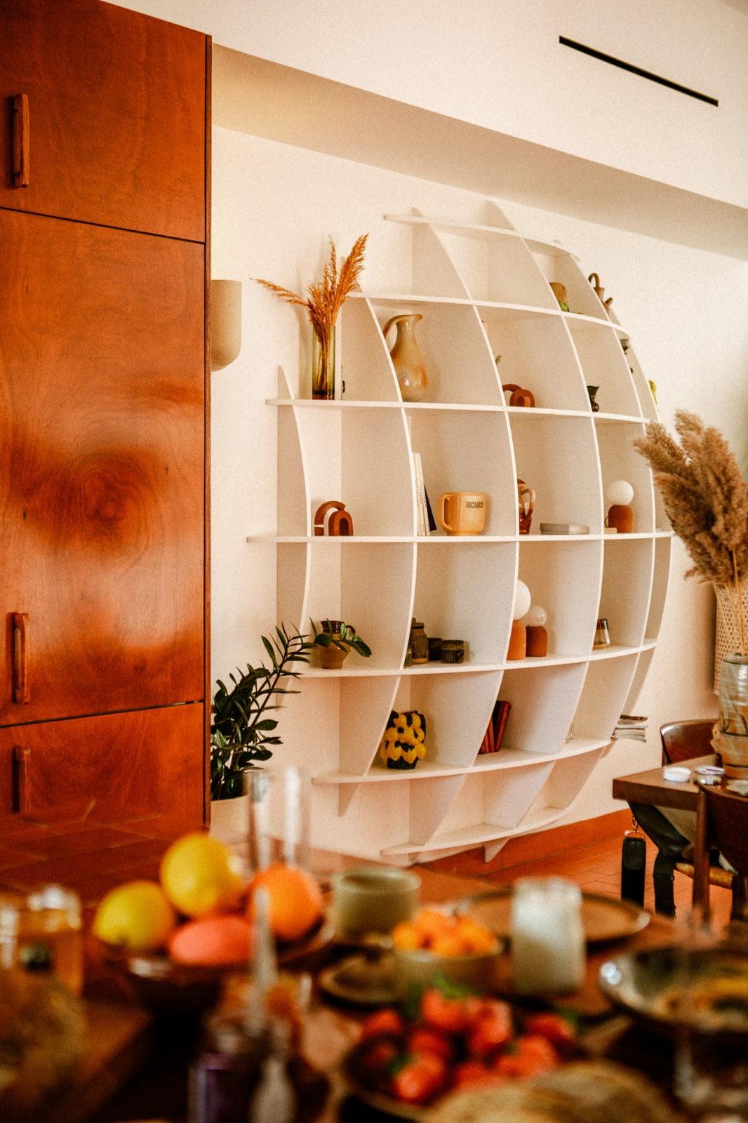 A cozy and modern living space with a unique white oval bookshelf filled with various decorative items, plants, and ceramics. To the left is a wooden Domino Deux cabinet, and in the foreground, a table with a variety of dishes and drinks. Pampas grass adds a natural touch.