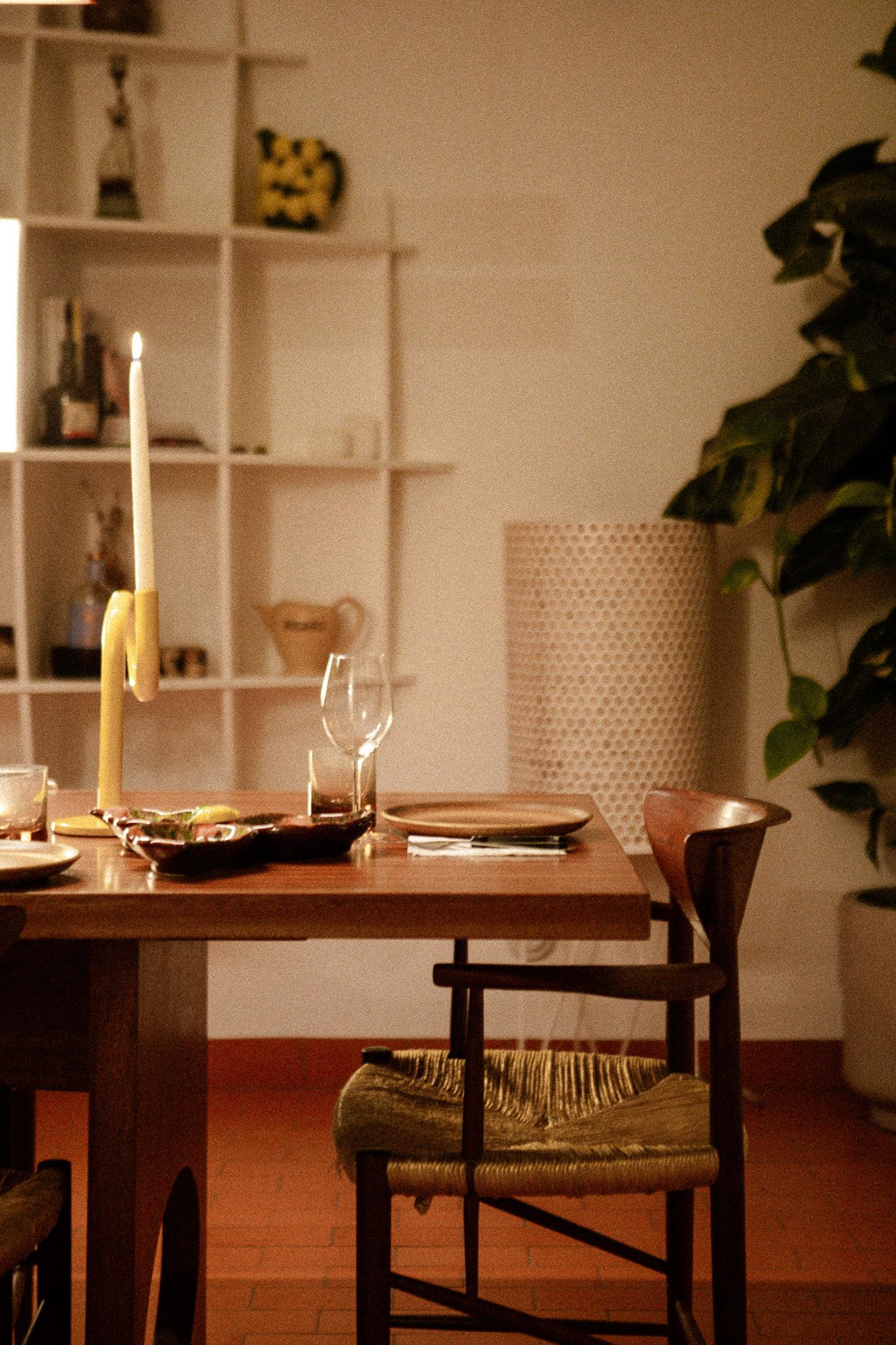 A cozy dining room with a wooden table and chairs, featuring a lit candle, cutlery, wine glasses, and a platter of food. In the background, there is a clean white shelf with decorative items, a large green plant, and a textured white lamp.