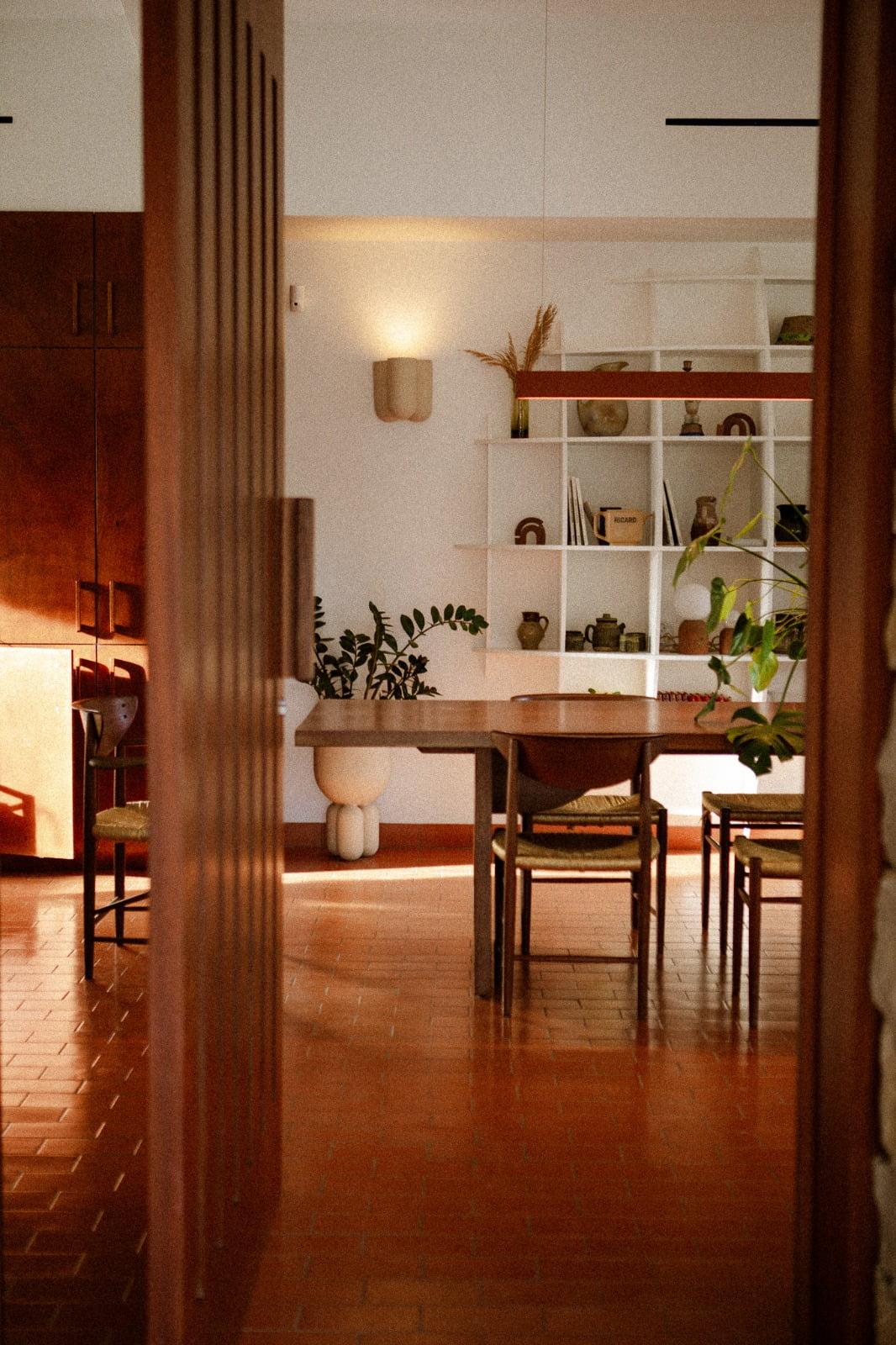 A cozy dining room with a wooden table surrounded by four chairs. The room is bathed in warm natural light and features a terracotta tiled floor. A white shelf on the back wall displays various decorative items, while several green plants add life to the space. The arrangement looks straight out of a Domino Deux magazine.