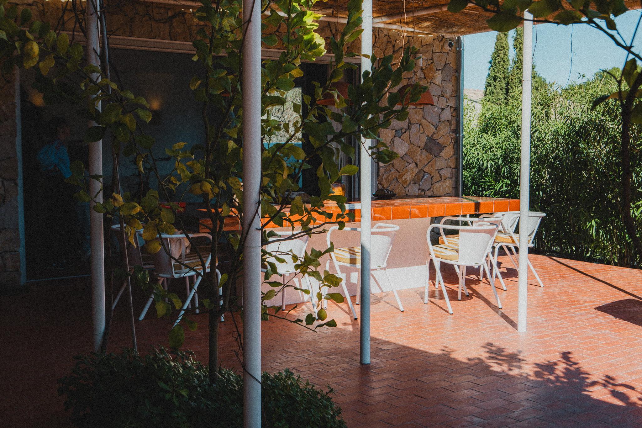 Outdoor terrace with a wooden pergola casting shadows over a dining area. The space includes white chairs around a long orange table, in front of a stone building. Green leafy plants and trees surround the terrace, creating a serene atmosphere.