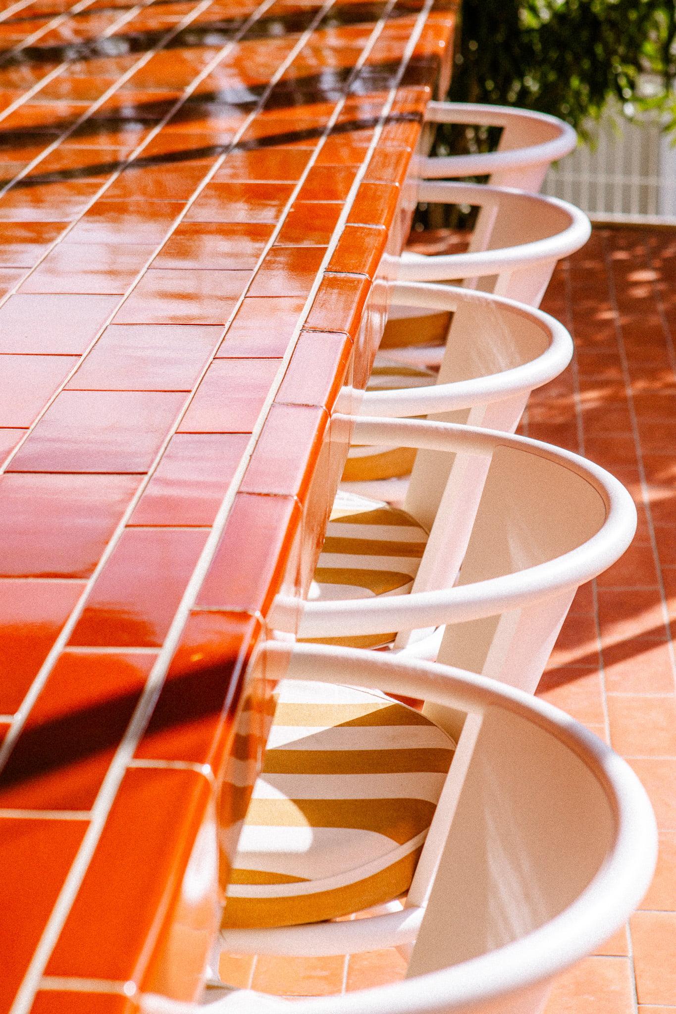 A row of modern bar stools, each featuring a white metal frame with circular backs and striped cushions, is lined up against a long bar counter tiled with glossy red tiles. Sunlight casts shadows on the setup, creating a bright and welcoming outdoor atmosphere.