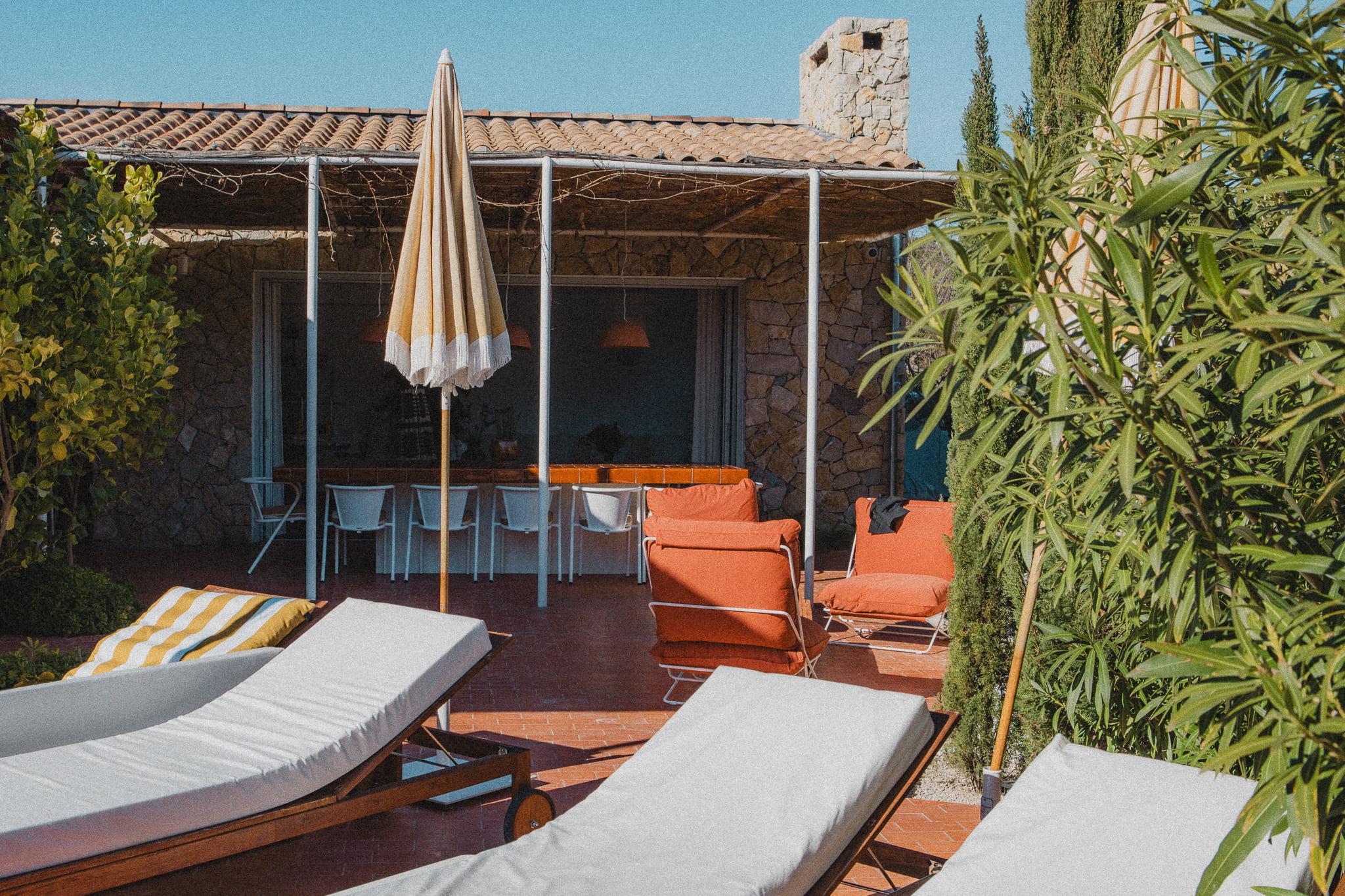 A cozy outdoor patio with lounge chairs and orange chairs under a large striped umbrella. A rustic stone house with a shaded dining area is in the background. Lush vegetation surrounds the space, adding to its inviting atmosphere.