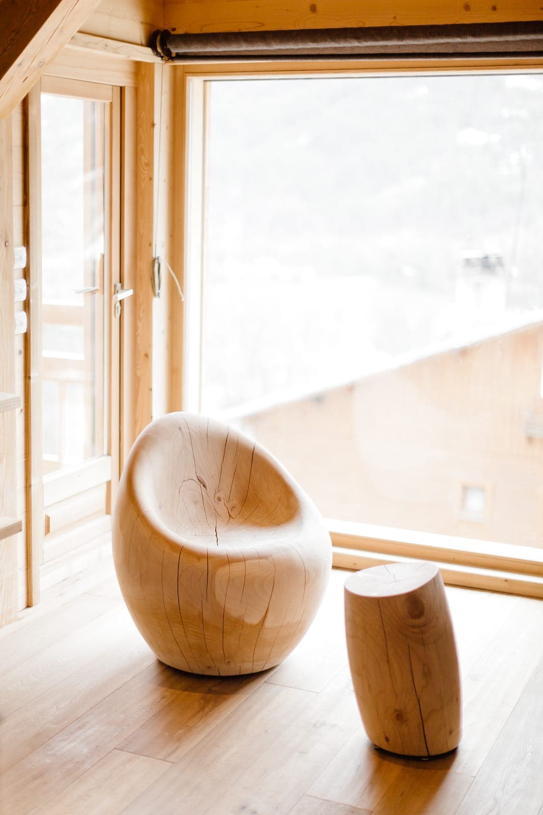 Une pièce chaleureuse avec de grandes fenêtres qui laissent entrevoir un paysage enneigé. À l'intérieur, une chaise en bois courbée unique et un tabouret en bois assorti sont posés sur un plancher en bois clair, créant une atmosphère chaleureuse et minimaliste.