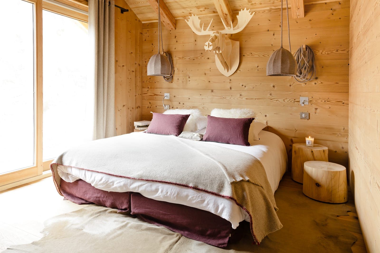 Chambre cosy à l'esthétique boisée avec un lit double et une literie blanche et bordeaux. Une tête d'élan en bois décore le mur au-dessus du lit. L'éclairage doux des lampes suspendues crée une ambiance chaleureuse et la lumière naturelle filtre à travers de grandes fenêtres.