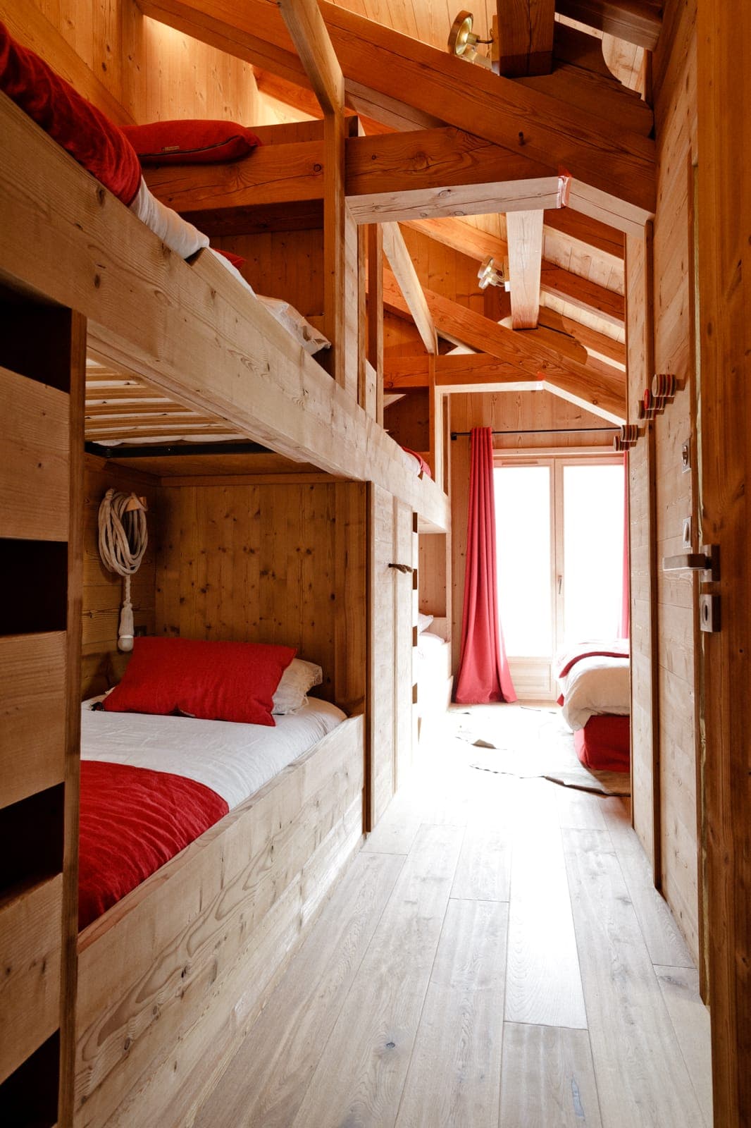 Intérieur de cabine en bois confortable avec lits superposés intégrés avec literie rouge et blanche. La lumière du soleil traverse une fenêtre avec des rideaux rouges, illuminant les poutres en bois et le sol, créant une atmosphère chaleureuse et accueillante.