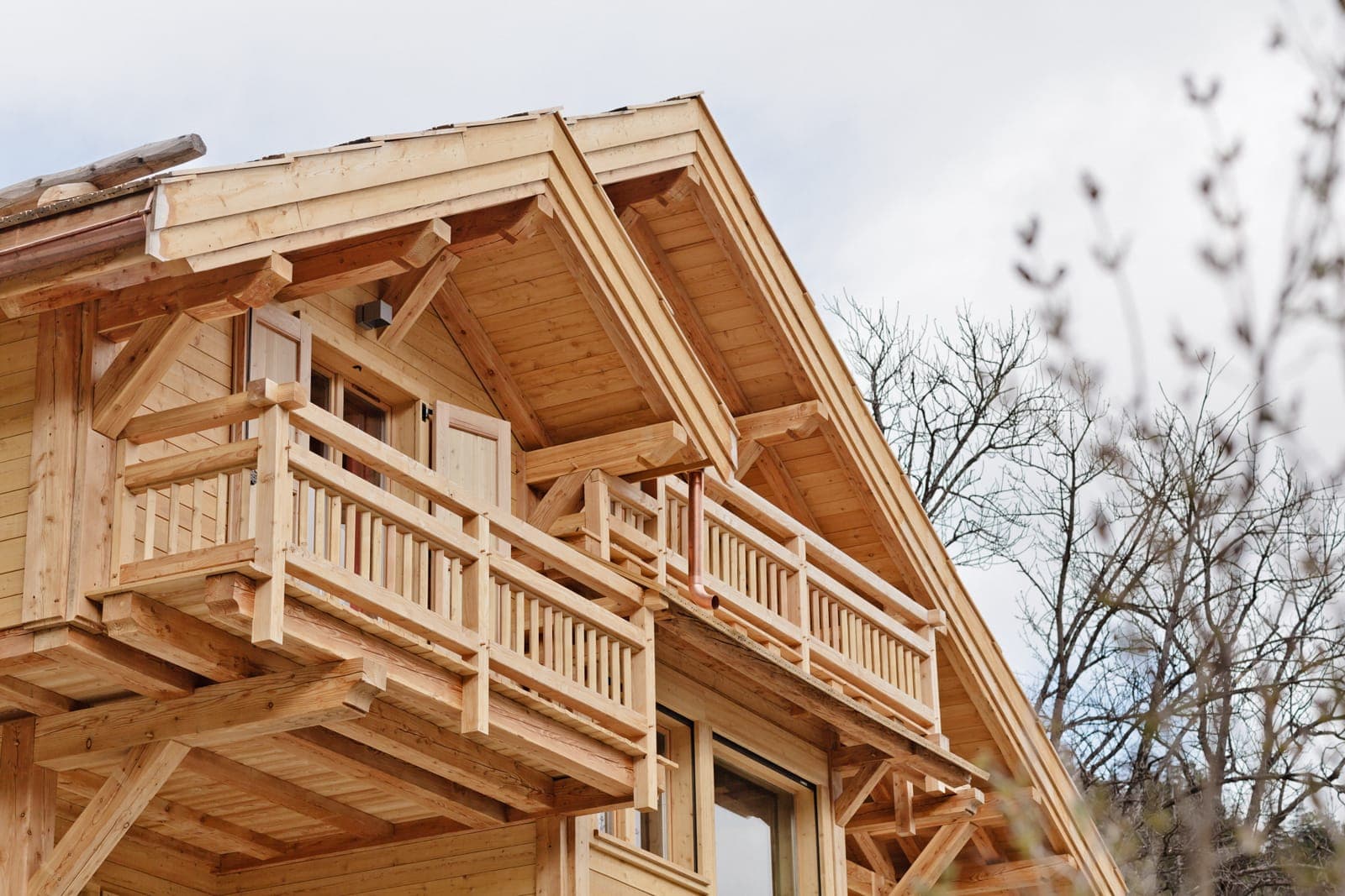 A wooden chalet with exposed beams and two balconies, viewed from below against a cloudy sky. Bare tree branches are visible on the right.