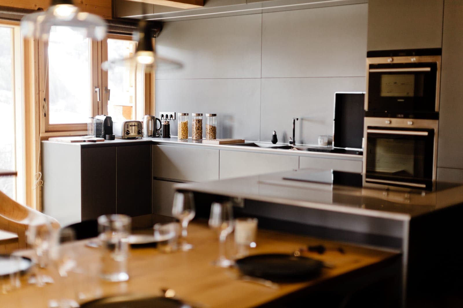 Cuisine moderne avec des armoires grises épurées, des fours muraux doubles et des appareils de comptoir, dont un grille-pain et une cafetière. Une table à manger au premier plan est dressée avec des assiettes et des verres. La lumière naturelle entre par une fenêtre.