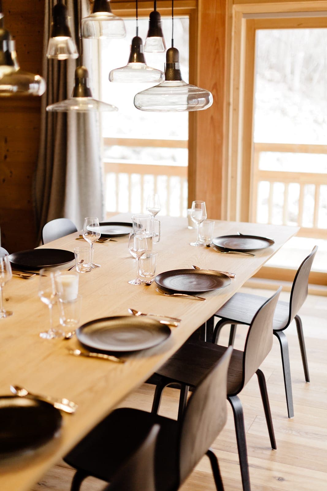 Une salle à manger moderne comprend une table en bois dressée avec des assiettes noires, des verres à vin et des couverts dorés. Des chaises noires élégantes entourent la table et de grandes fenêtres ajoutent de la lumière naturelle. Des suspensions de style industriel sont suspendues au-dessus.