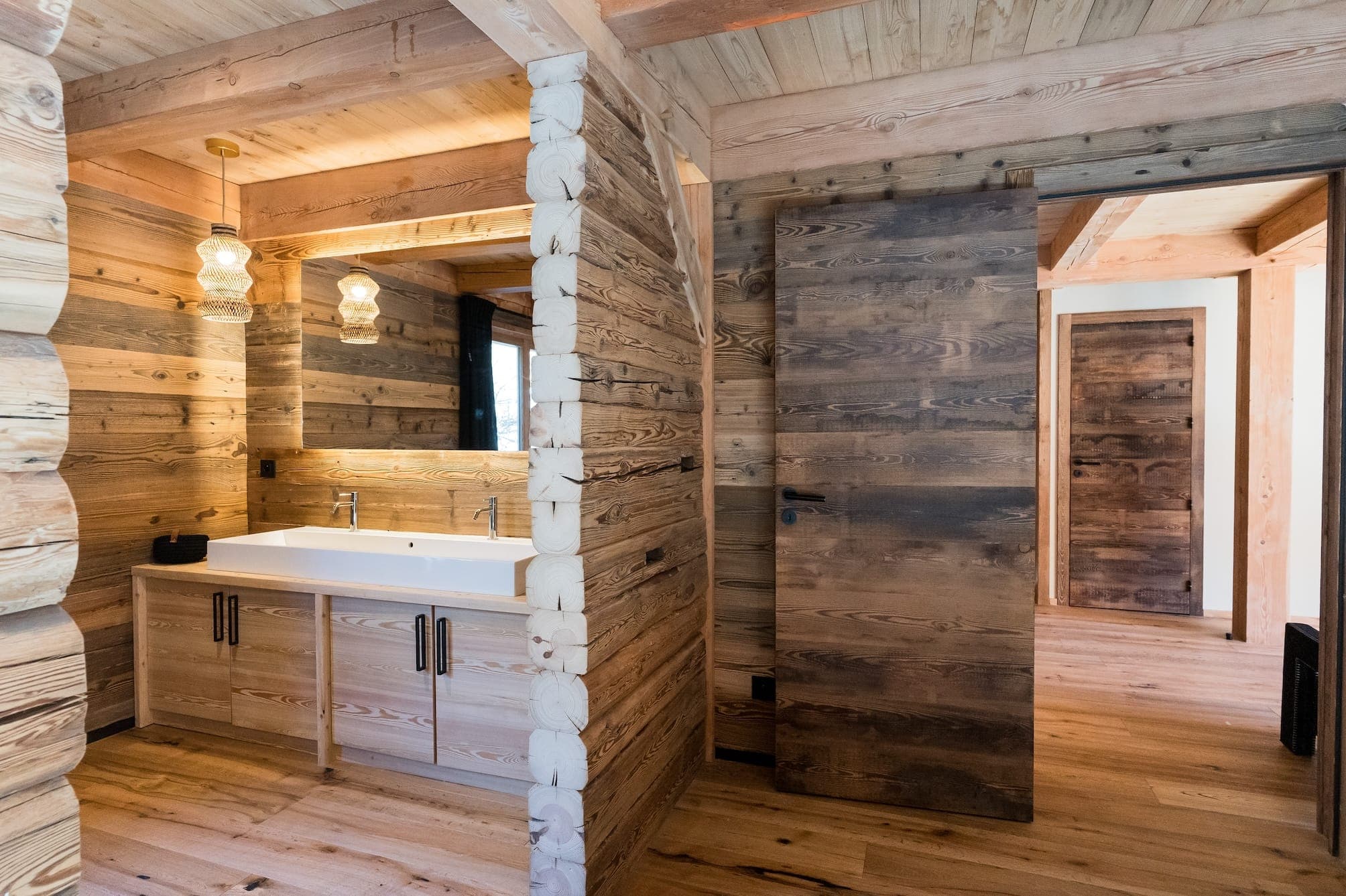 Un intérieur chaleureux en bois avec une salle de bain avec un meuble à double vasque et deux luminaires suspendus. La salle de bain communique avec un couloir aux murs et au sol en bois, menant à une porte fermée. L'espace dégage une atmosphère rustique et naturelle.