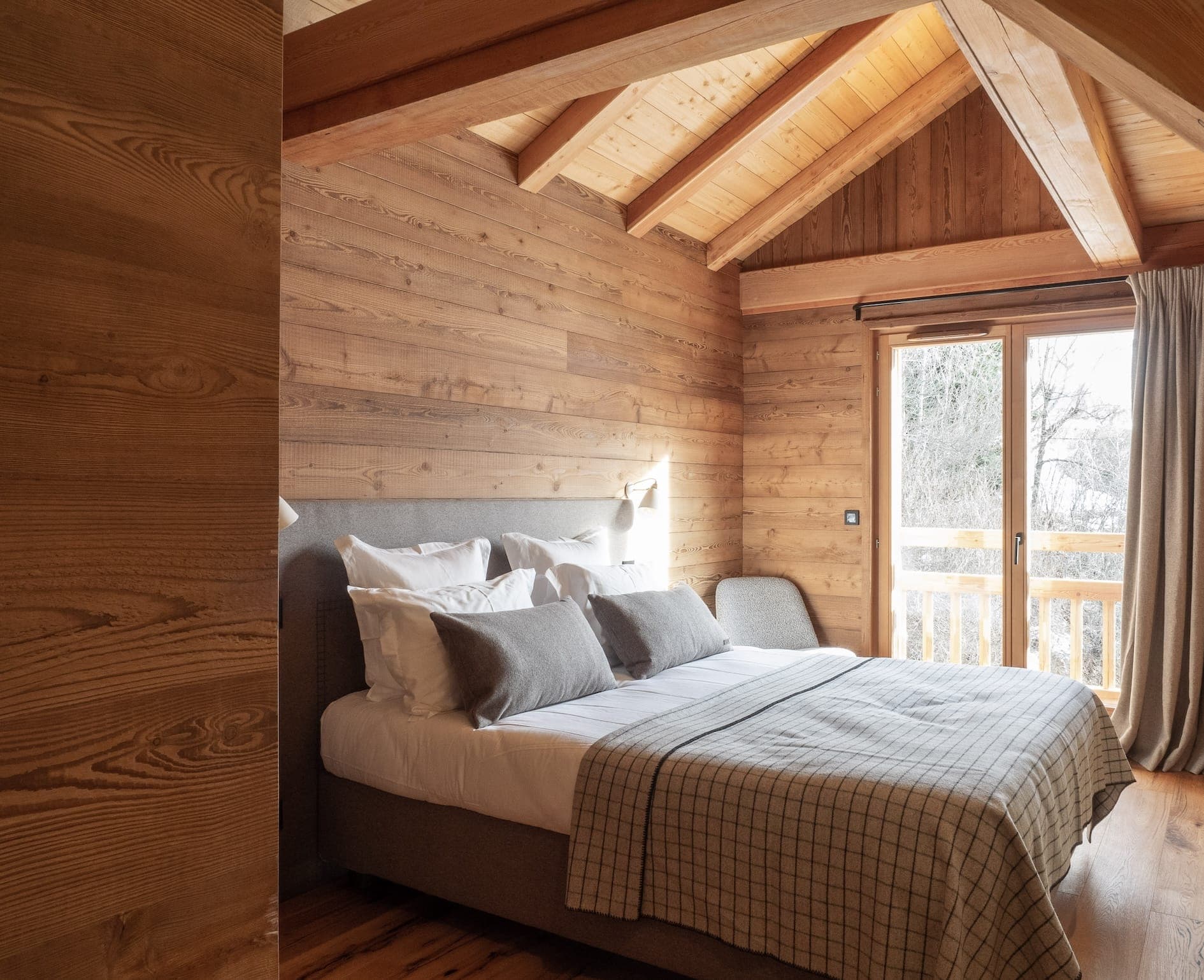 Une chambre confortable avec des murs et un plafond lambrissés, dotée d'un grand lit avec une literie blanche et grise. La lumière du soleil filtre à travers une porte vitrée menant à un balcon avec vue sur les arbres. La pièce dégage une atmosphère chaleureuse et accueillante.