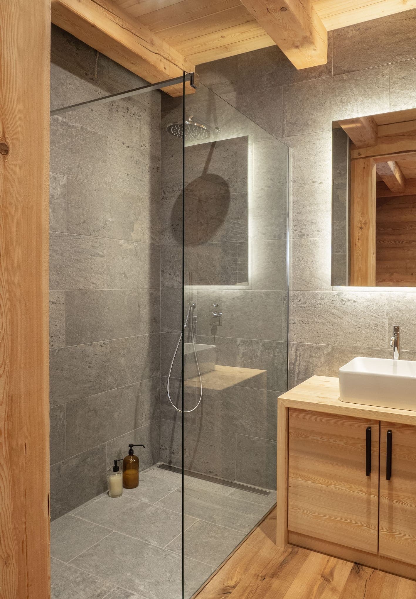 Salle de bains moderne avec une grande douche vitrée et des carreaux de pierre grise. Une vanité en bois avec un lavabo rectangulaire blanc et des accessoires noirs complète le plafond et le sol en bois rustique. Un miroir rétroéclairé est suspendu au-dessus du lavabo.