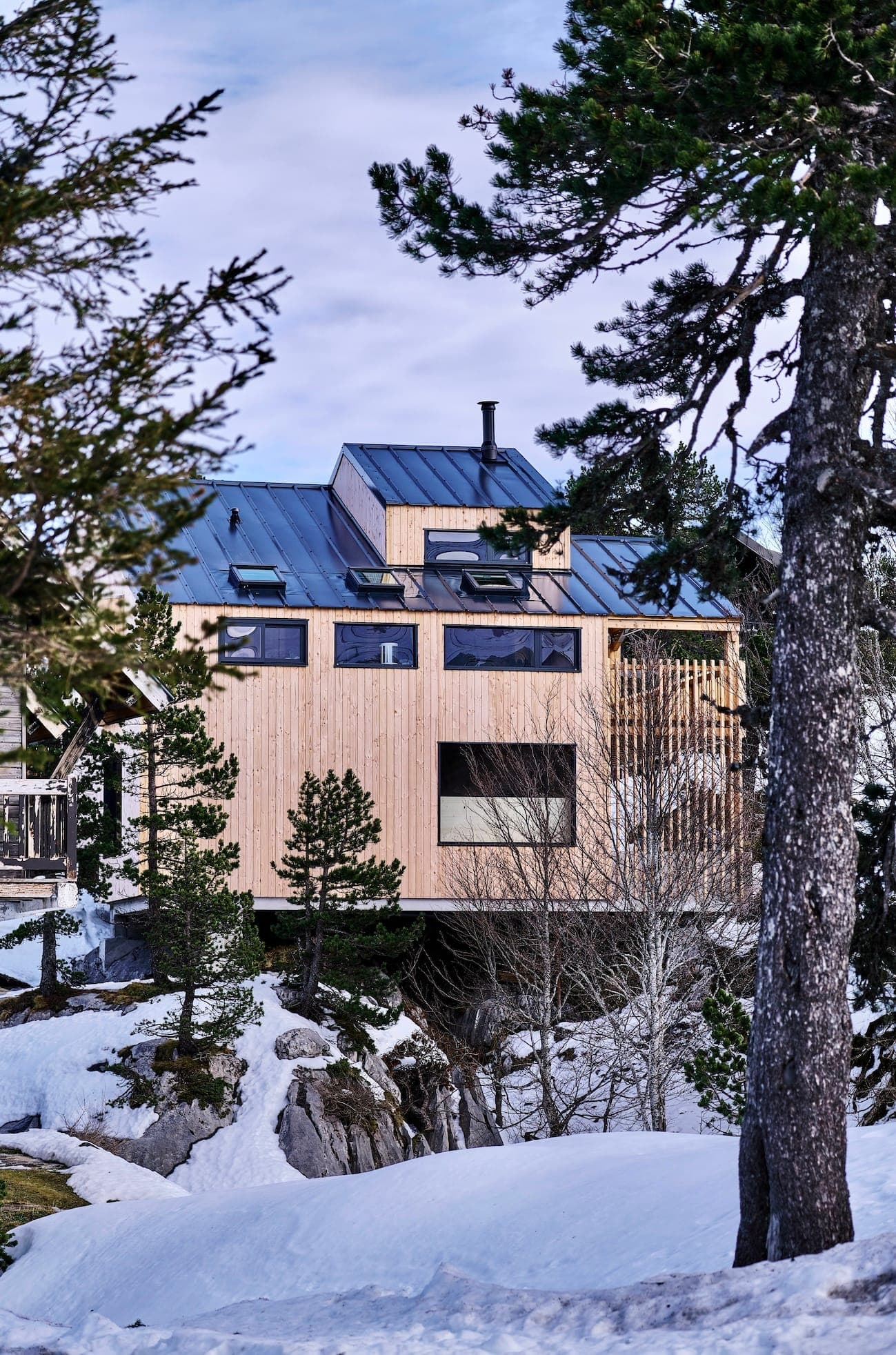 Une cabane moderne en bois avec de grandes fenêtres et un toit en métal, entourée de sol enneigé et de pins, sous un ciel partiellement nuageux.