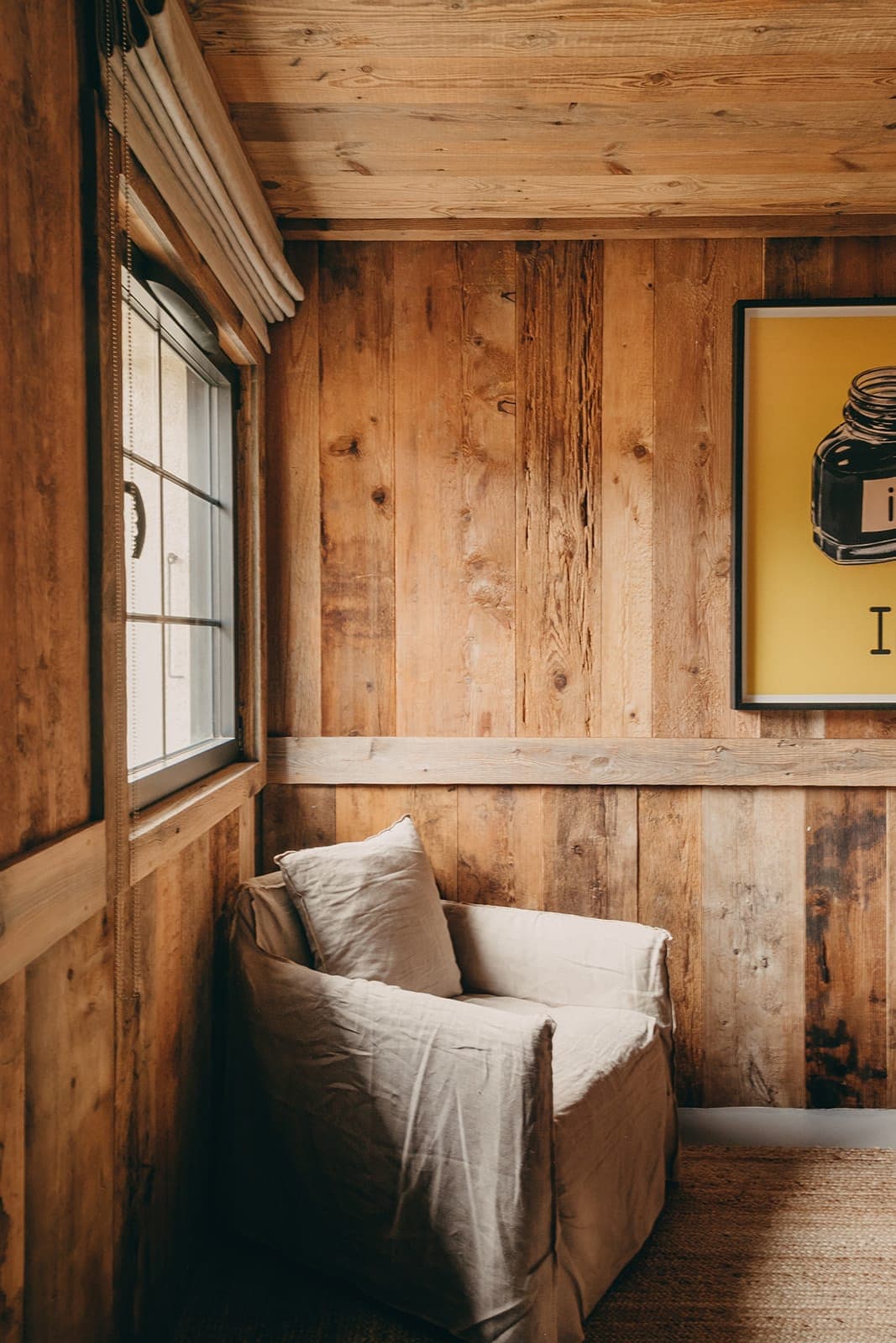 A cozy room with wooden walls and ceiling features a light gray cushioned armchair. A window with closed blinds lets in soft light. A colorful framed artwork is hung on the wall beside the chair.