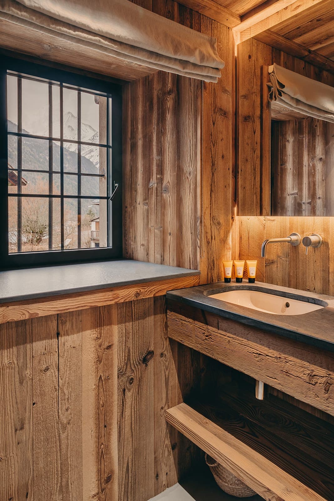 Rustic bathroom with wooden walls and a window view of snow-capped mountains. Features include a stone sink, modern faucet, small toiletries, and a mirror. Warm lighting highlights the natural wood textures and cozy atmosphere.