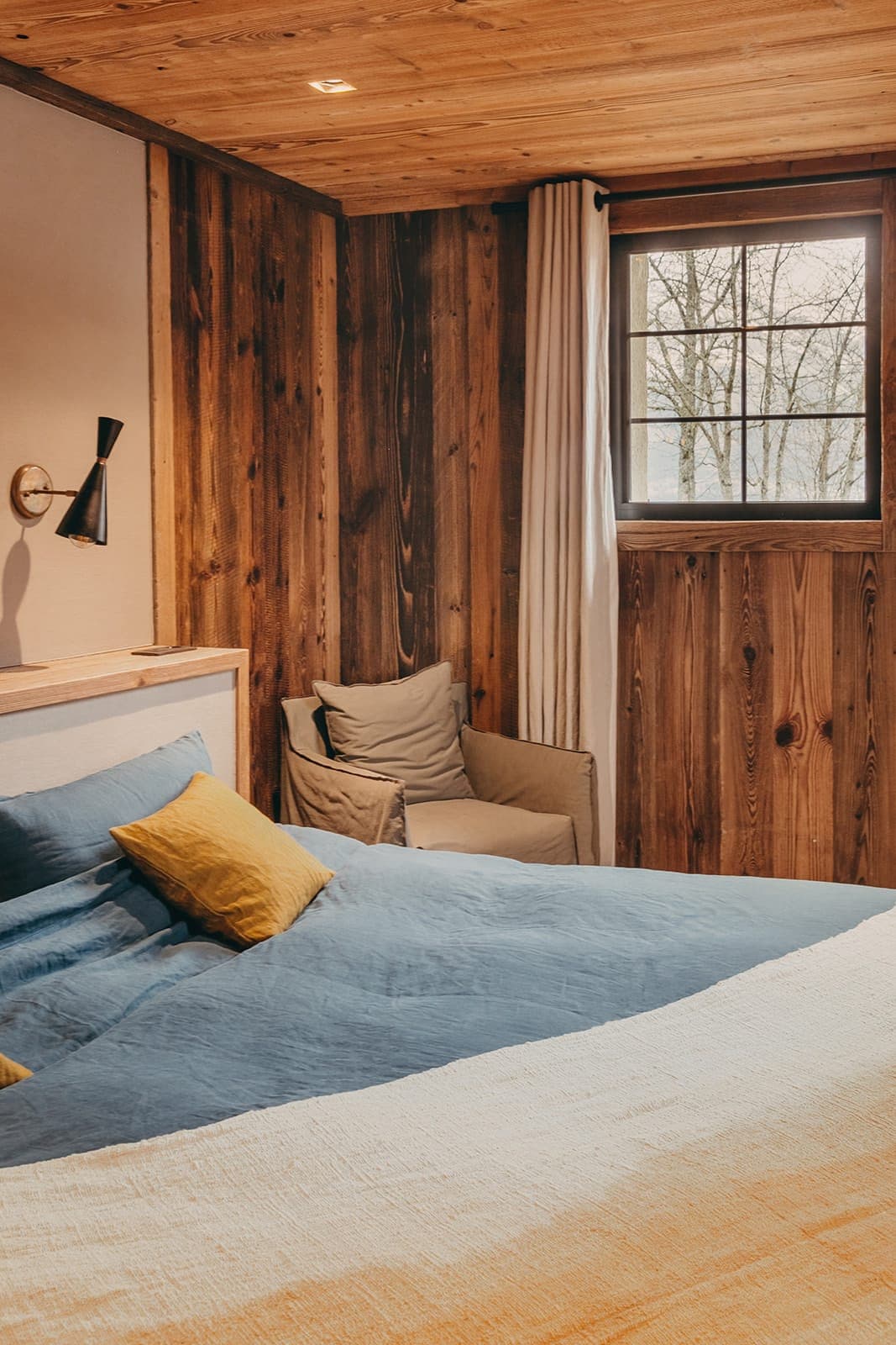 A cozy bedroom with wooden walls and a large window. The bed has blue and yellow bedding, with a matching yellow pillow. A cushioned chair is in the corner, and a wall-mounted lamp provides warm lighting. A curtain hangs beside the window.