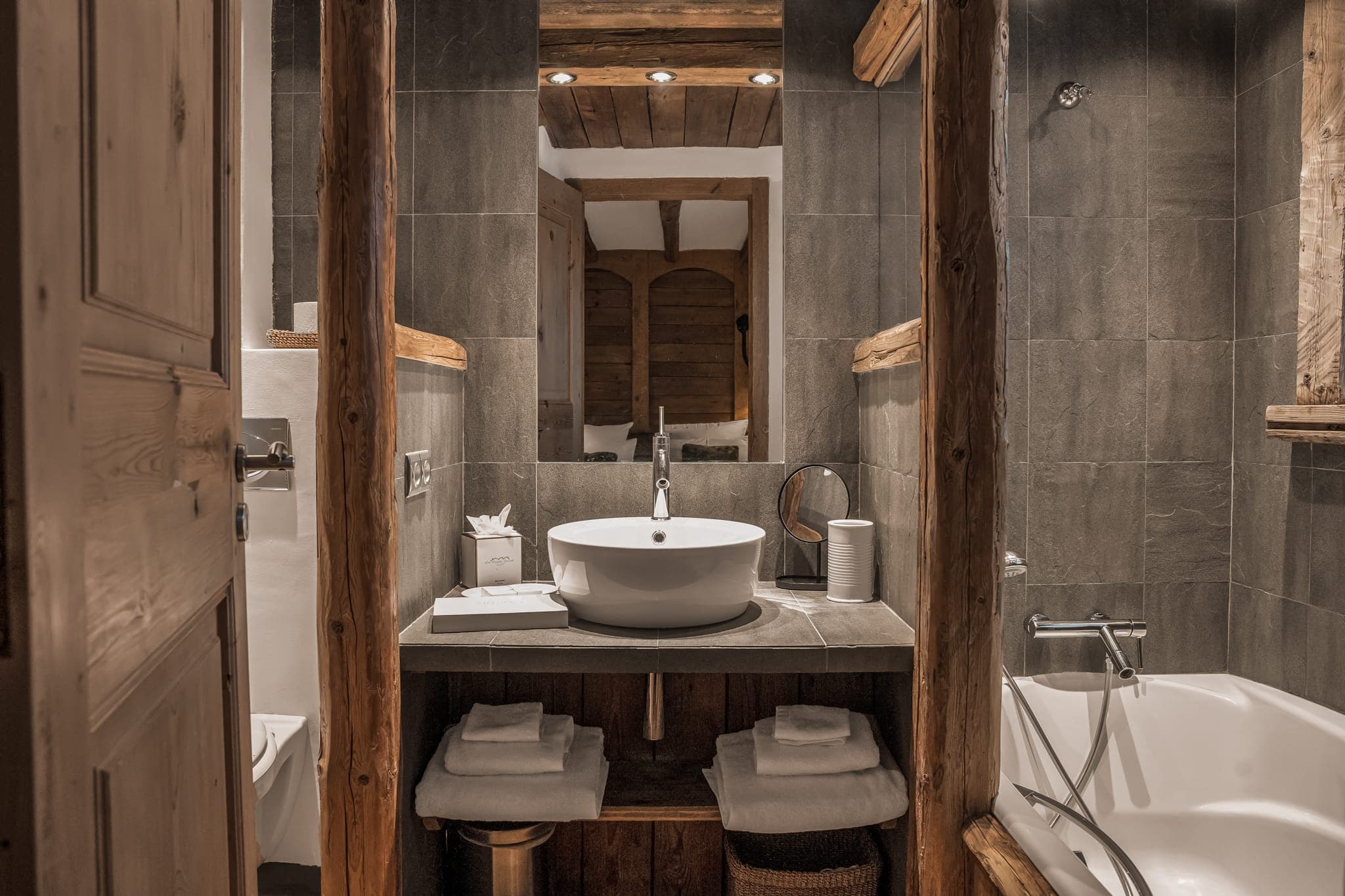 A cozy bathroom with rustic wooden accents features a modern white sink atop a wooden vanity, surrounded by dark grey tiled walls. Towels are neatly stacked on shelves below the sink, and a tub is situated to the right side of the image. Soft lighting adds warmth.