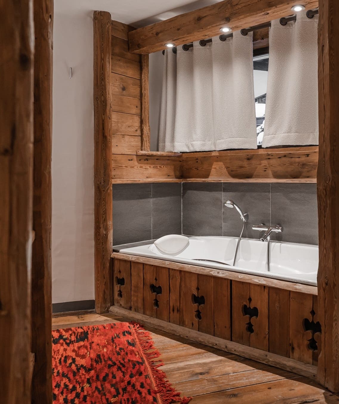 A rustic bathroom features a wooden-framed bathtub with decorative cutouts, gray tile backsplash, and an overhead white curtain. A red and black patterned rug lies on the wooden floor. The walls and ceiling are paneled with natural wood.