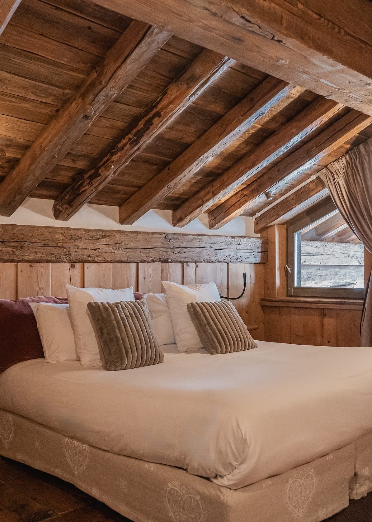 A cozy wooden attic bedroom featuring a bed with white linens adorned with several pillows and cushions. The room has a rustic feel with exposed wooden beams on the ceiling and a small window letting in natural light, partially covered by beige curtains.