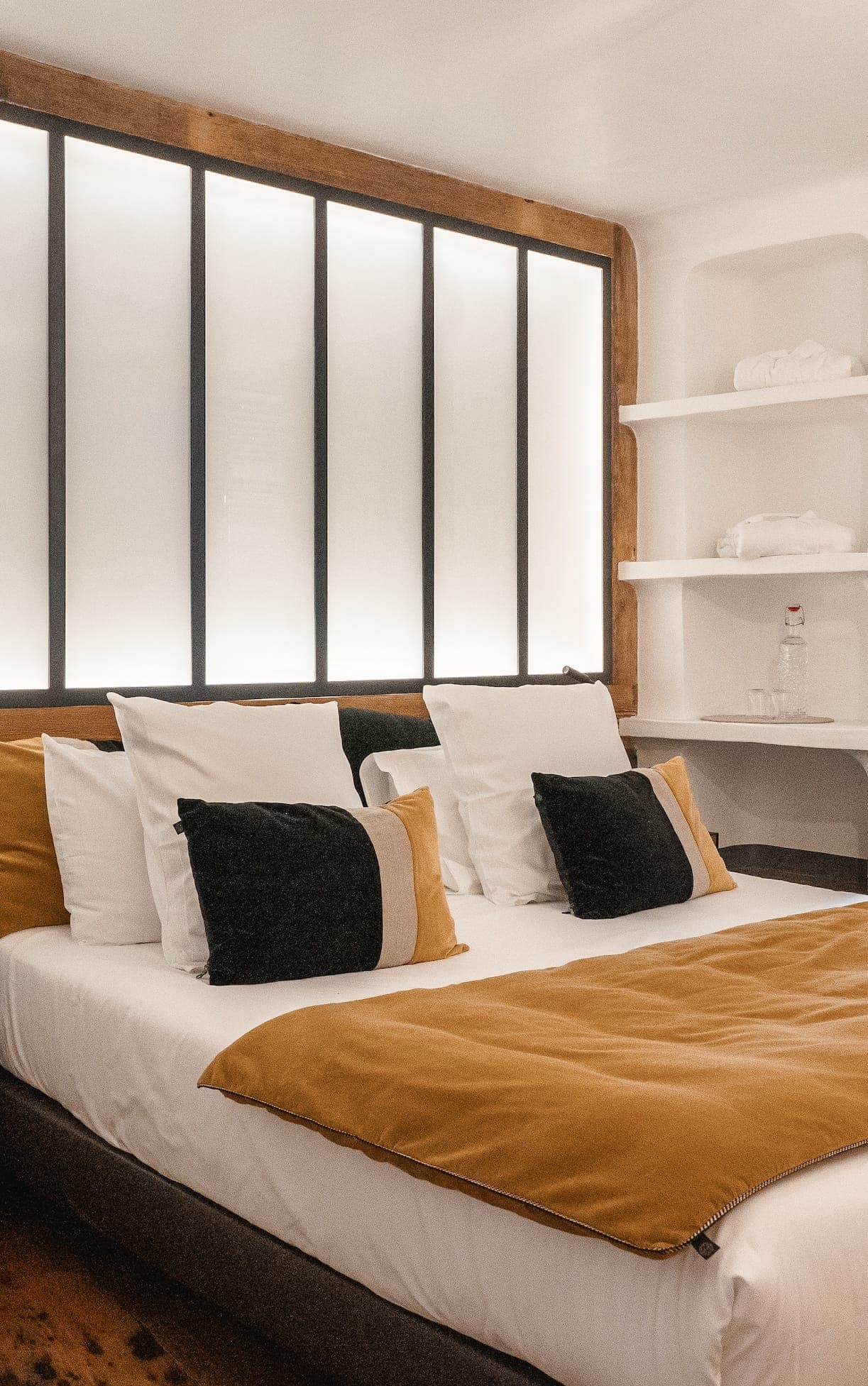 A modern bedroom features a neatly made bed with white and mustard-colored bedding, along with several black and white pillows. The room has a large, backlit headboard and built-in shelving on the right, holding white towels and a bottle of water.