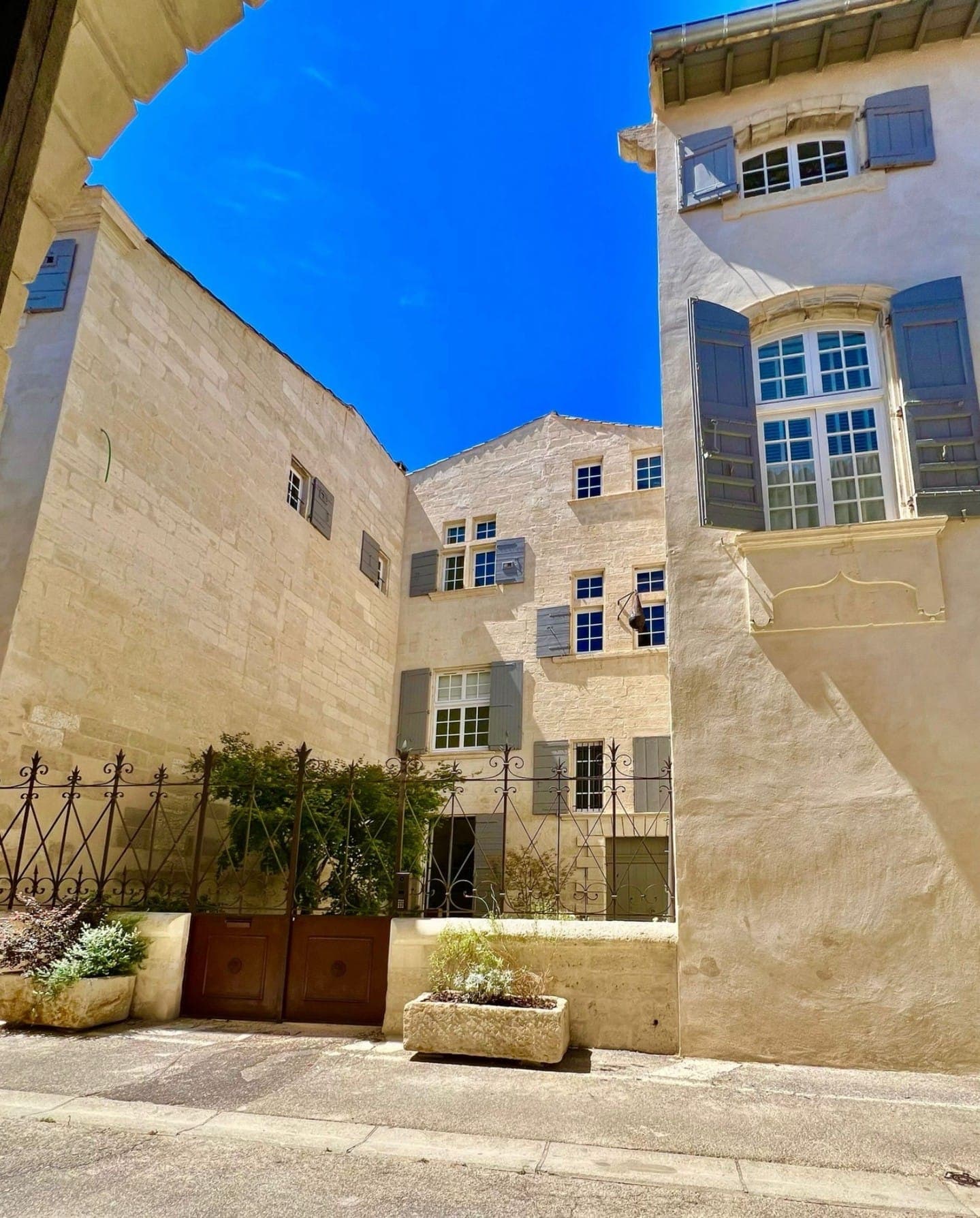 Une cour ensoleillée entourée de bâtiments en pierre beige avec des volets bleus et de multiples fenêtres. Le ciel est clair et d'un bleu vif. Une clôture métallique ornée et quelques jardinières avec de la verdure sont visibles au premier plan.