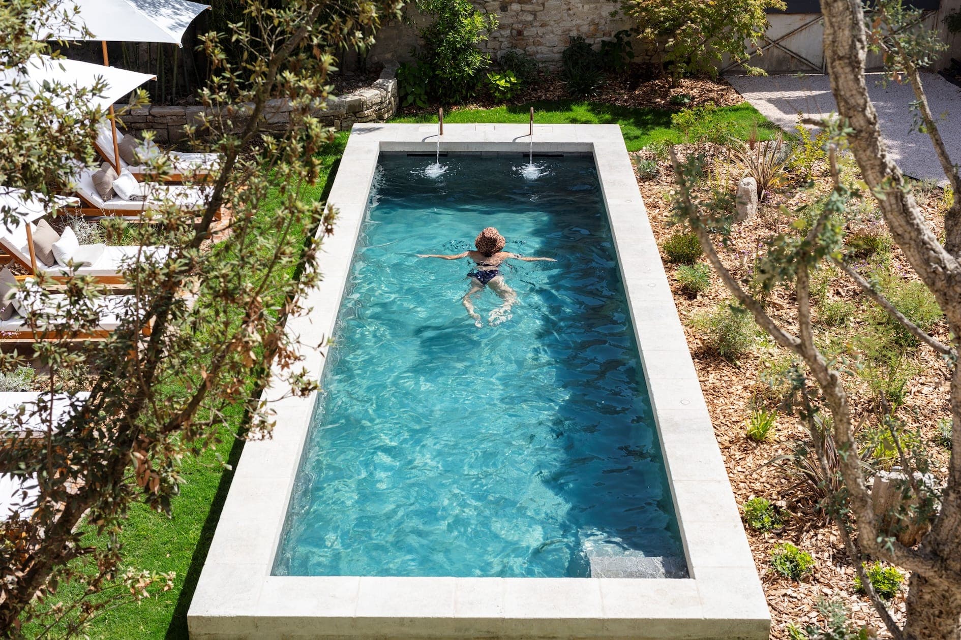 Une personne nage tranquillement dans une piscine rectangulaire étroite entourée d'une végétation luxuriante et de chaises longues. L'eau est claire et ensoleillée, créant un cadre extérieur tranquille.