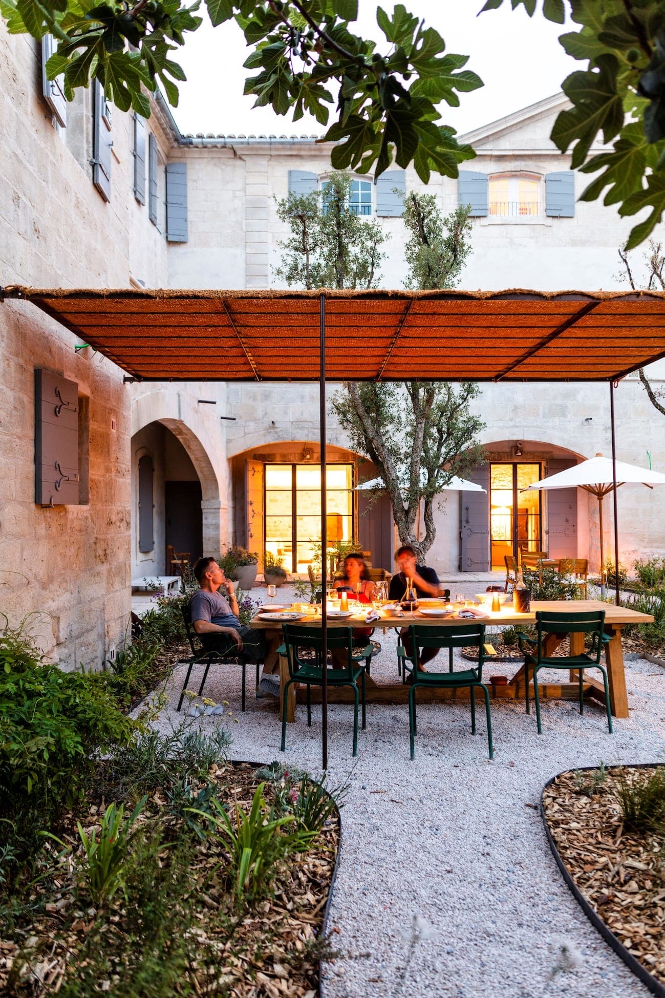 Une cour extérieure confortable avec une table à manger avec plusieurs personnes assises, chaleureusement éclairée par des bougies. L'espace est entouré de verdure et flanqué d'un bâtiment en pierre avec des volets et des arcades en bois. Un auvent tissé fournit de l'ombre au-dessus de la table.