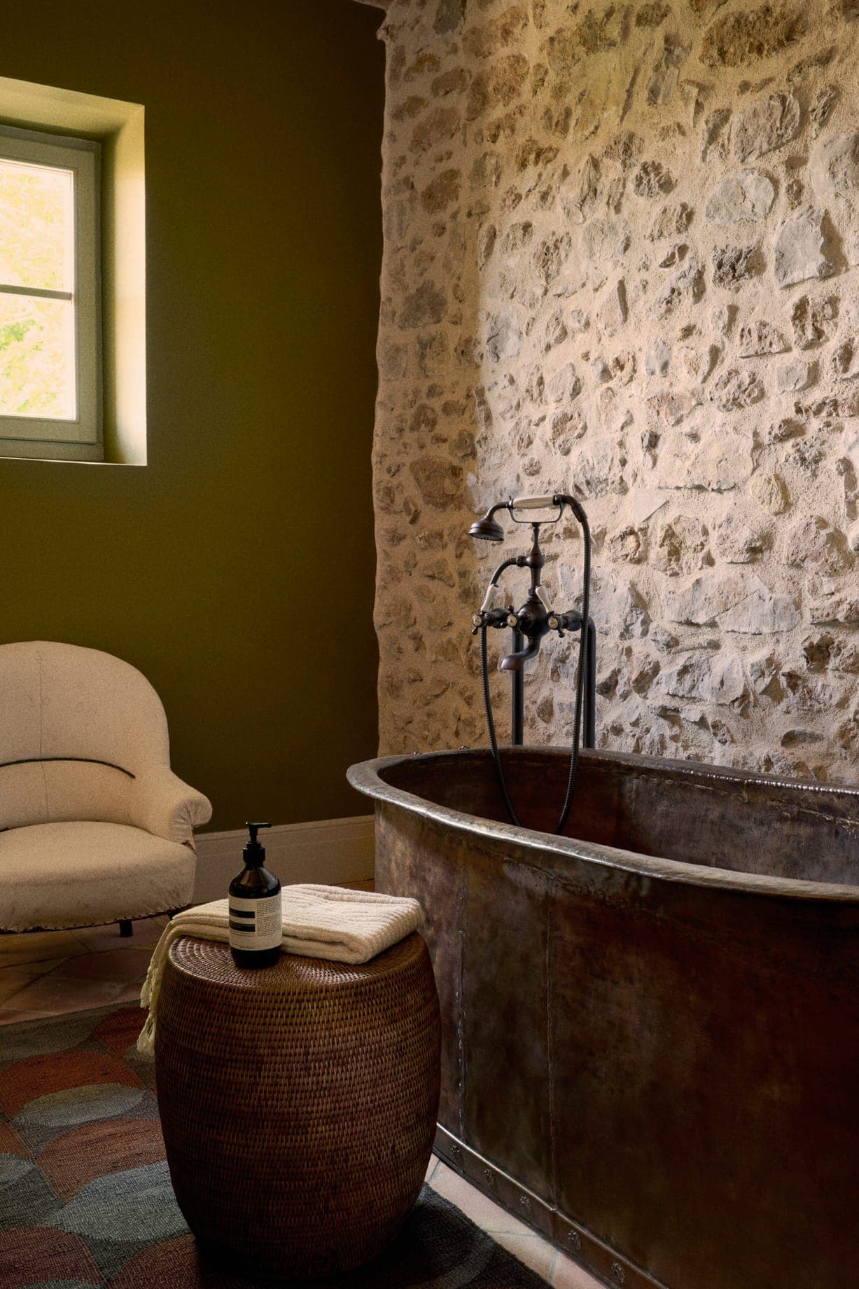 Une salle de bain confortable est dotée d'une baignoire en métal rustique contre un mur en pierre texturé. Un fauteuil beige est placé à côté d'un tabouret tressé, qui contient une serviette et un distributeur. Une petite fenêtre laisse entrer la lumière naturelle, mettant en valeur les tons terreux de la pièce.