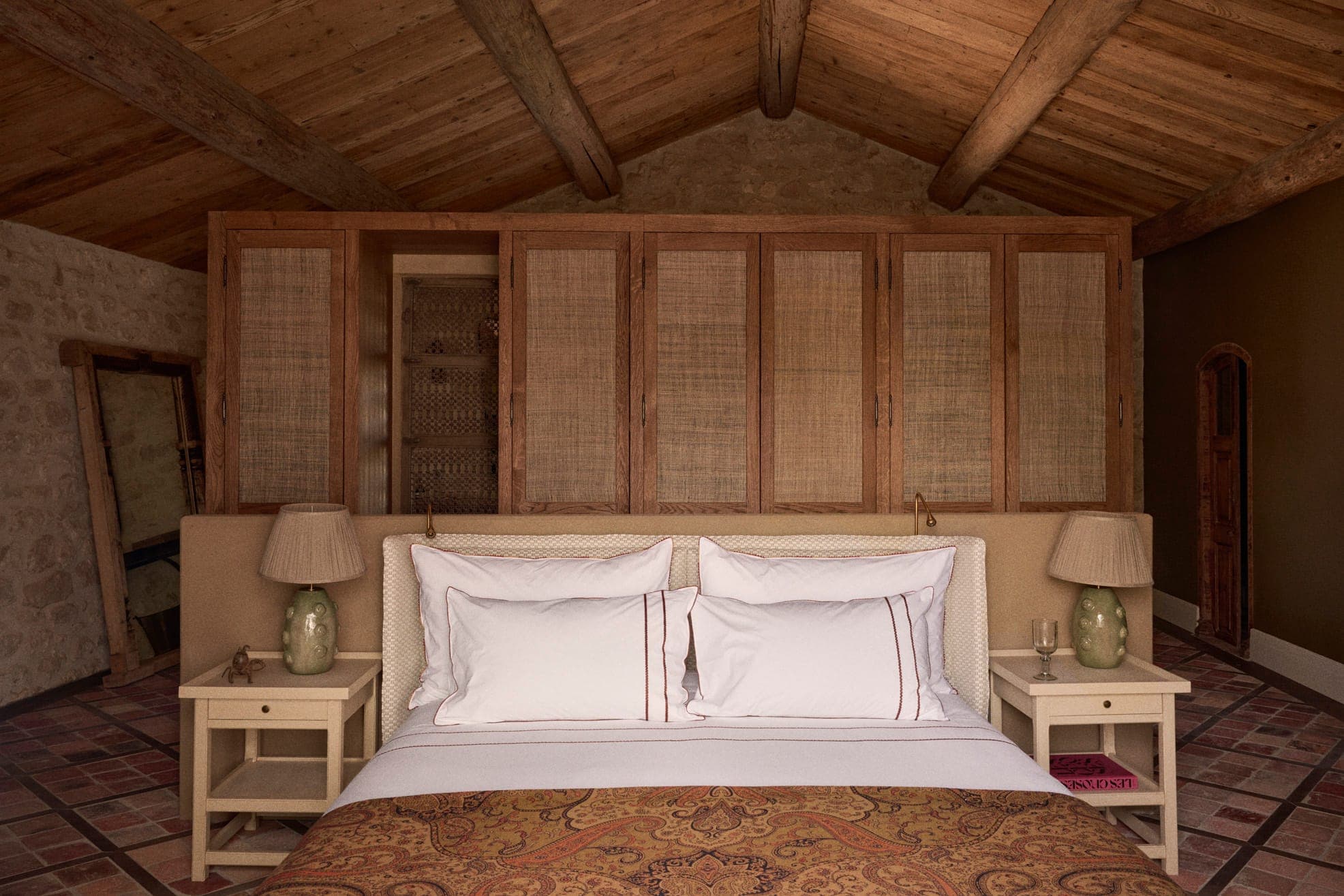 Une chambre confortable au décor rustique dispose d'un grand lit avec des oreillers blancs et une literie à motifs, flanqué de deux tables de chevet et de lampes. La pièce est dotée de poutres en bois au plafond, d'un mur en pierre et d'une cloison avec des panneaux tissés.