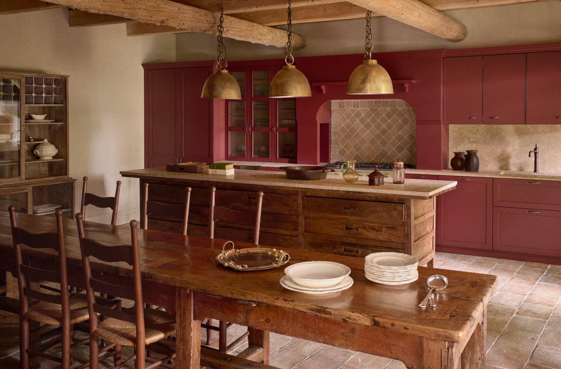 Rustic kitchen with wooden beams, red cabinets, and a weathered wooden table set with plates and a magnifying glass. Two large brass pendant lights hang over a wooden island with farmhouse elements and vintage decor throughout.