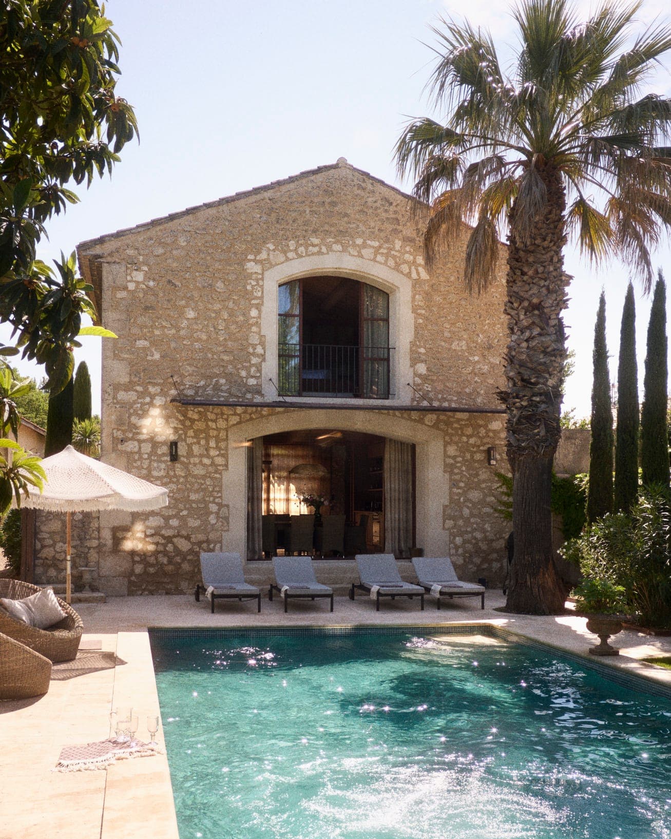 Une maison rustique en pierre avec des volets en bois et un balcon donne sur une piscine sereine. Quatre chaises longues sont disposées au bord de la piscine. Un grand palmier et des cyprès encadrent la maison, et un parasol blanc est visible sur la gauche.