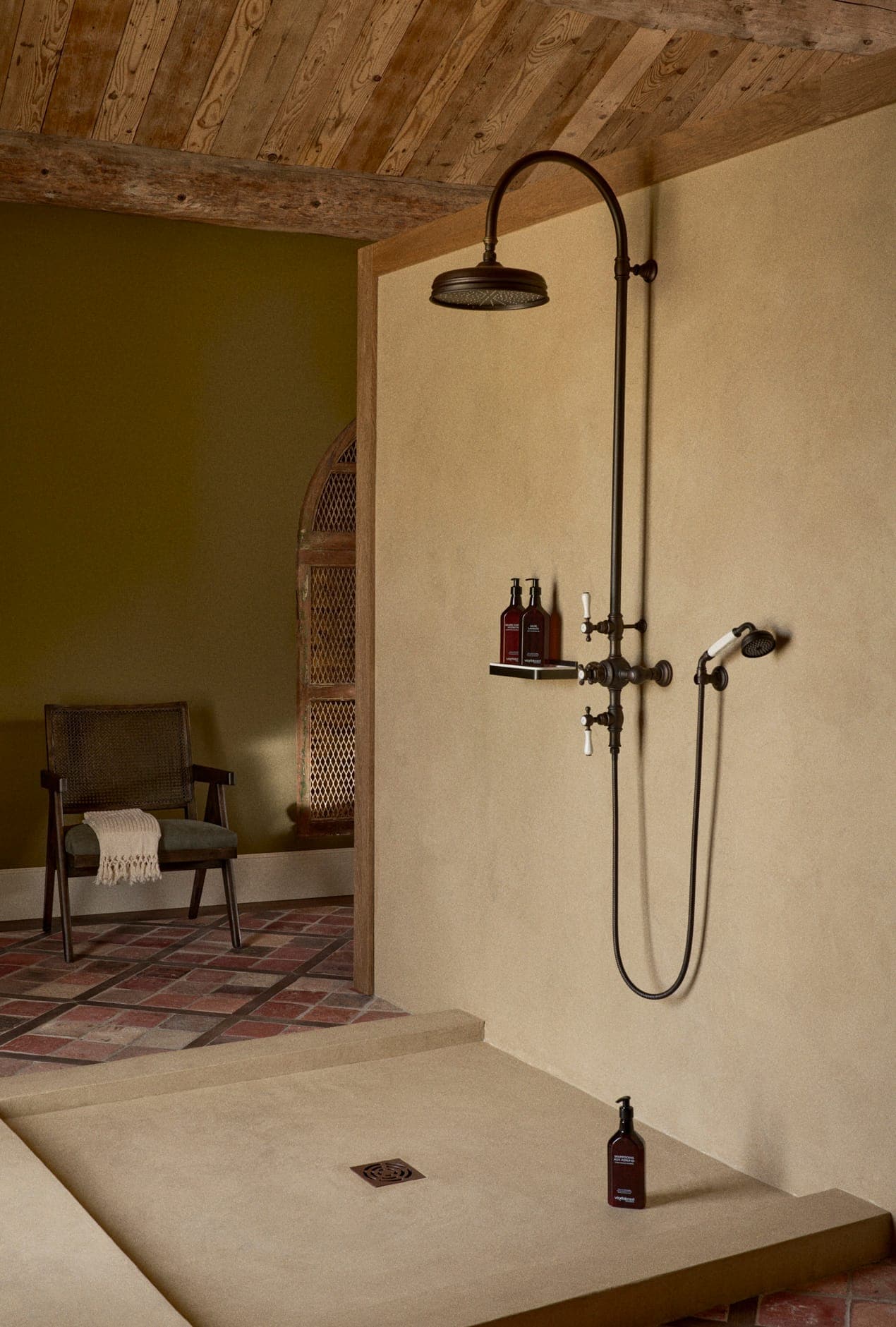 Un espace douche rustique avec une pomme de douche noire mate et des accessoires sur un mur beige. Des bouteilles de produits de toilette sont posées sur une petite étagère. Le sol est carrelé et une chaise recouverte d'une serviette trône à l'arrière-plan. Le plafond est en bois.