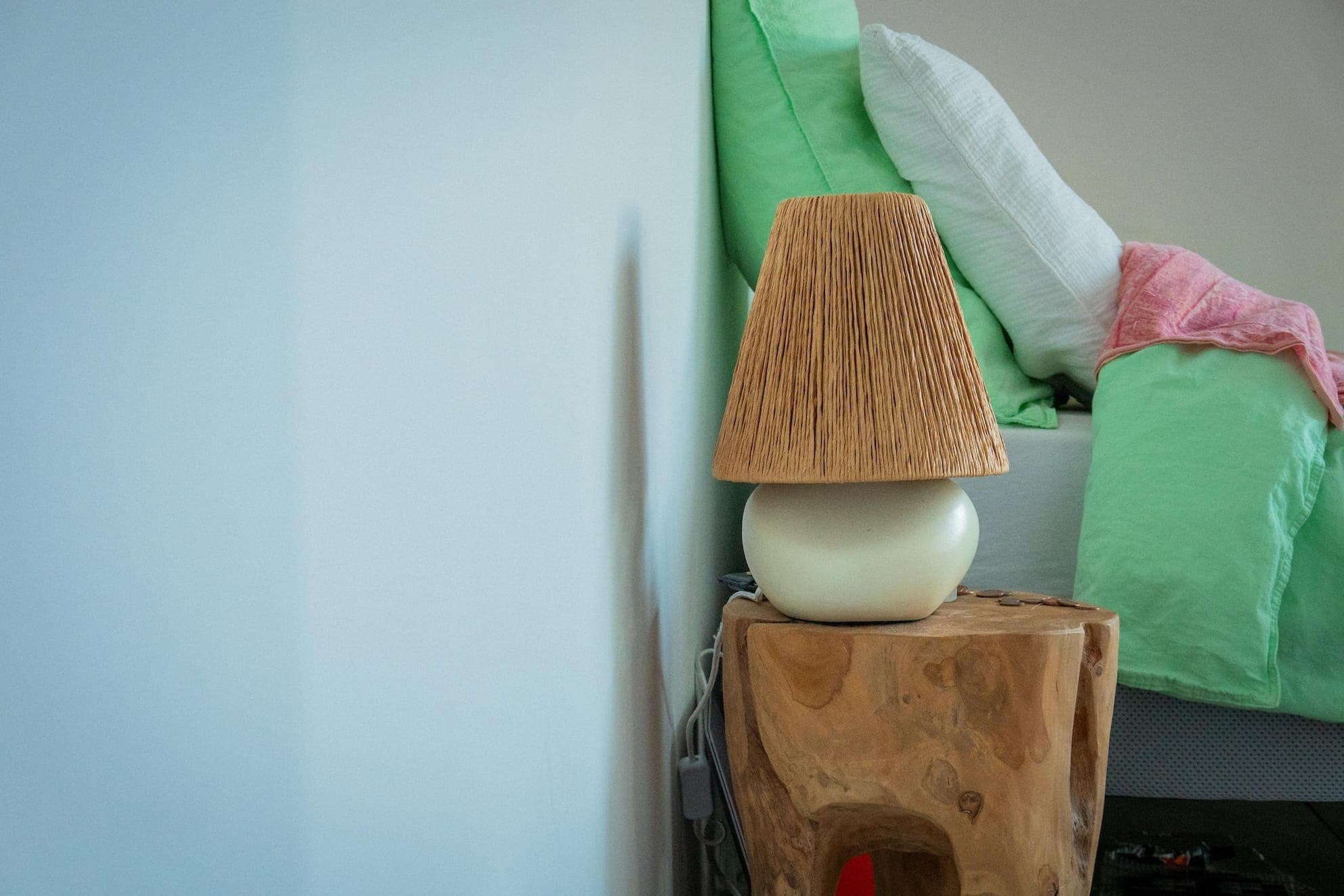 Une chambre minimaliste avec une table de chevet en bois et une lampe à abat-jour texturé. Le lit est orné d'une literie verte et blanche et d'un plaid rose, posé contre un mur de couleur claire.