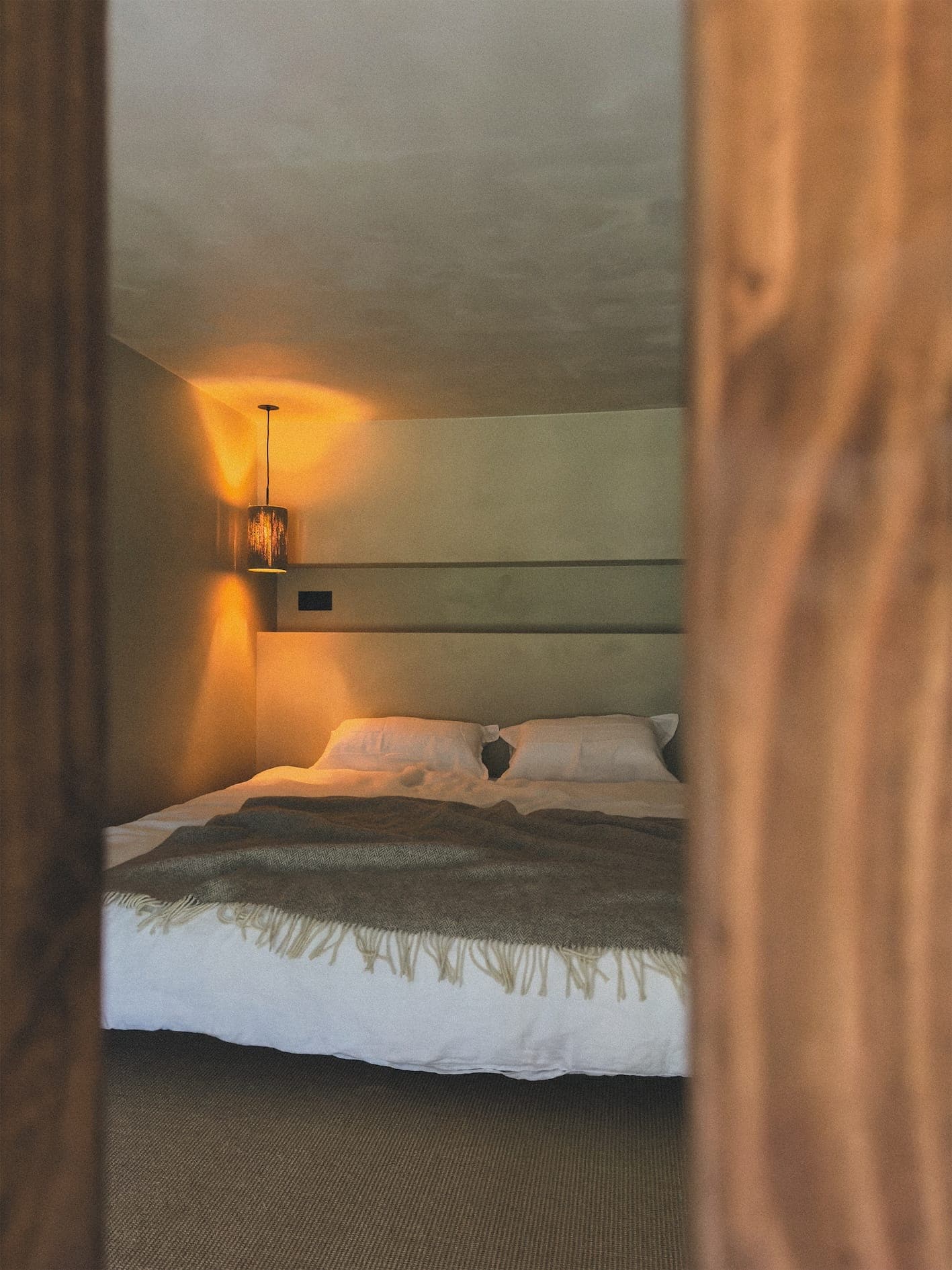 Une chambre confortable avec un éclairage chaleureux, avec un lit soigneusement fait avec des draps blancs et une couverture marron. Une douce lueur d'une lampe suspendue illumine l'espace, créant une atmosphère relaxante. Le bois encadre la vue sur la pièce.