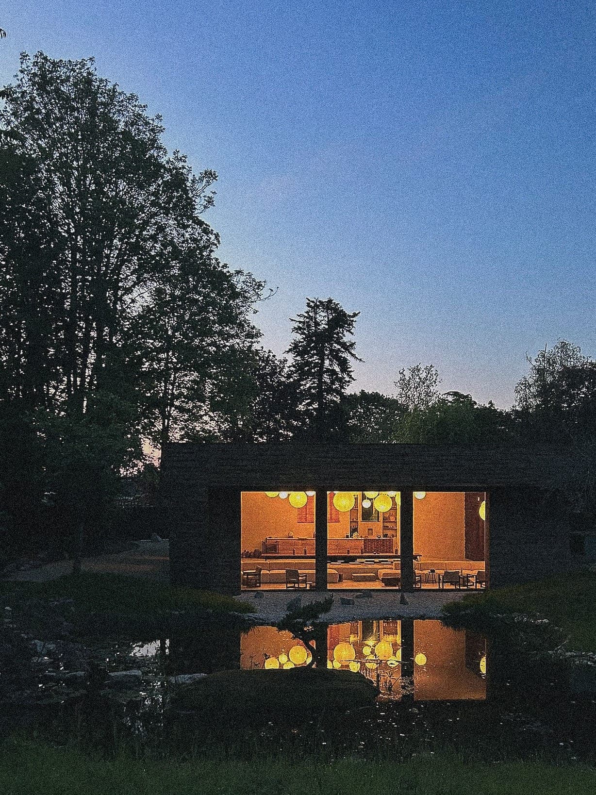 Au crépuscule, une cabane confortable et éclairée, dotée de grandes fenêtres, est entourée d'arbres. Les lumières chaleureuses de l'intérieur se reflètent dans un étang serein au premier plan, contrastant avec le ciel bleu profond du soir.