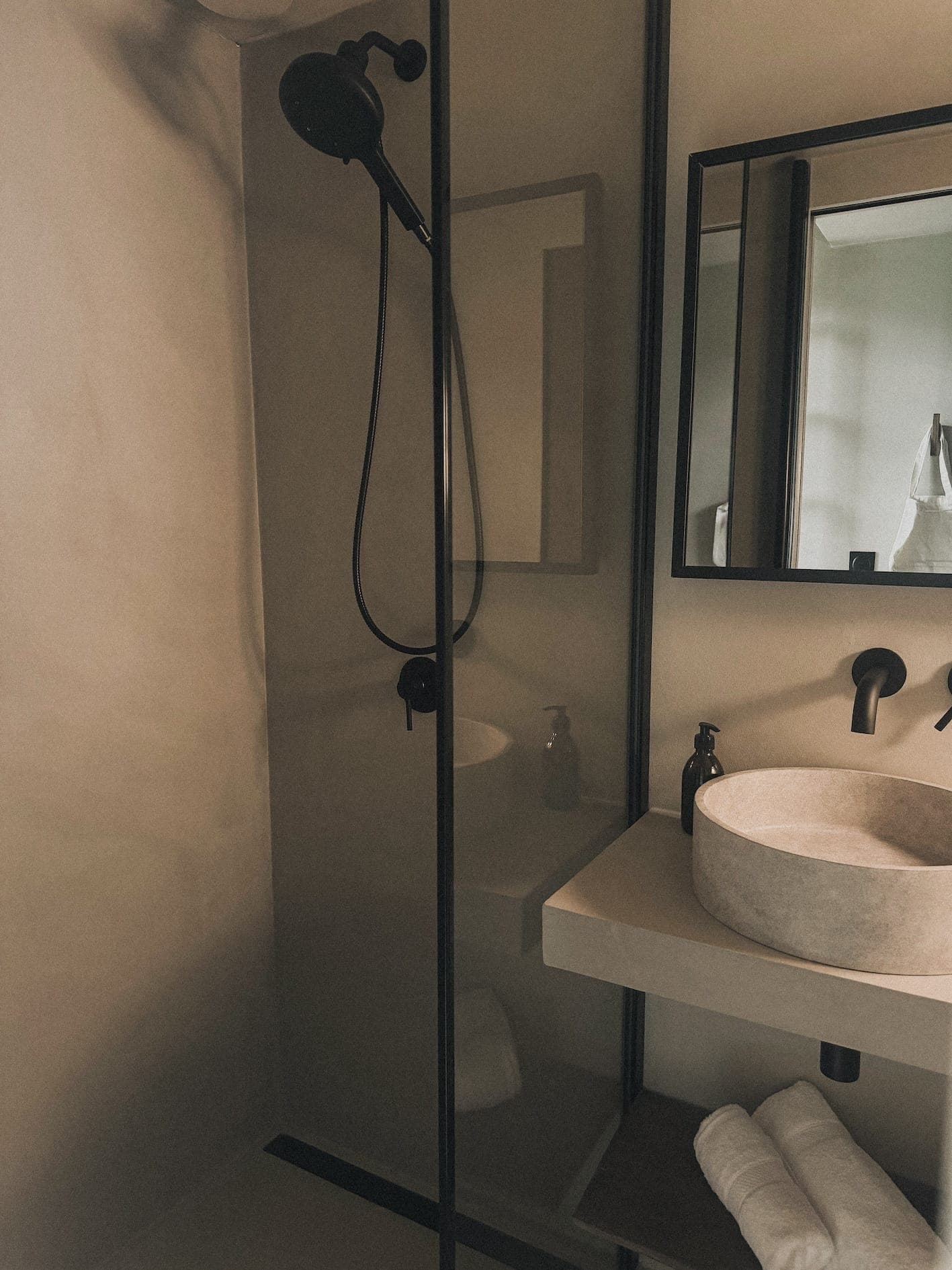 Salle de bains moderne avec un lavabo en pierre sur une vasque flottante, des accessoires noirs et un miroir. Une douche à l'italienne avec une cloison en verre se trouve sur la gauche. Des serviettes blanches roulées sont placées sous le lavabo.