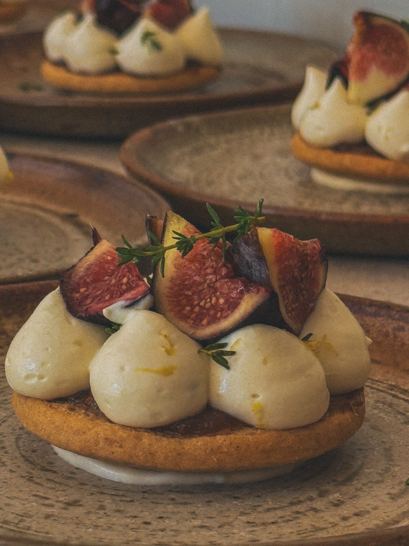 Un dessert composé d'une base de biscuits ronds garnie de pépites crémeuses et de tranches de figues fraîches, garnies de petites herbes vertes, disposées élégamment sur une assiette en céramique.