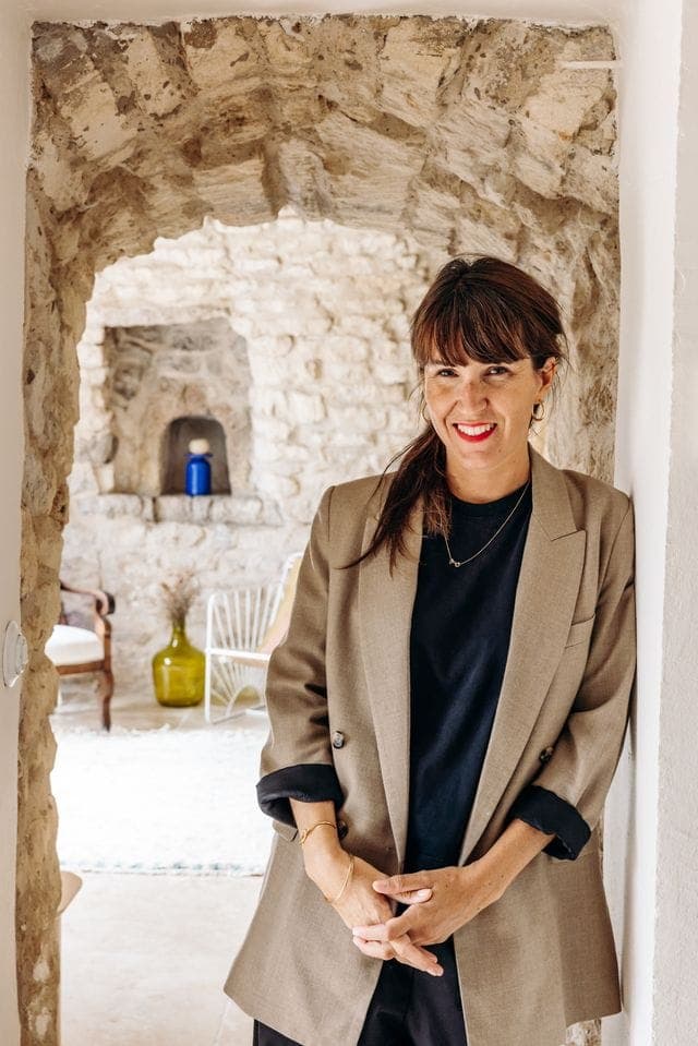 Une femme vêtue d'un blazer beige et d'une chemise noire se tient sous une arche en pierre rustique. Elle sourit, les bras croisés, et l'arrière-plan présente une pièce confortable aux murs en pierre avec une chaise blanche et des objets décoratifs.