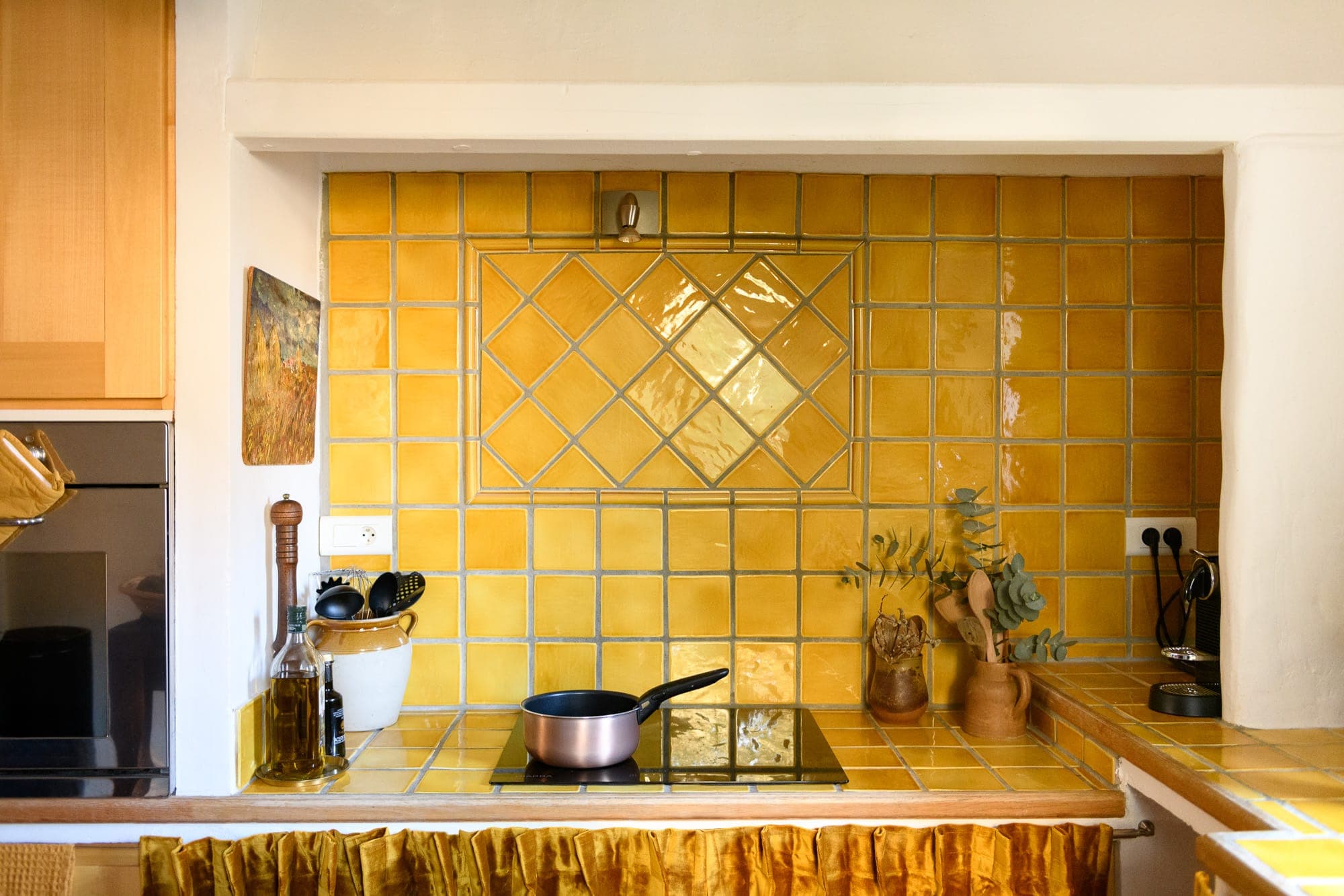 A kitchen features a vintage yellow tile backsplash and countertop, with a black stove top and pink saucepan. Wooden utensils, green foliage in a vase, and various jars and bottles are neatly arranged. A small painting decorates the wall.