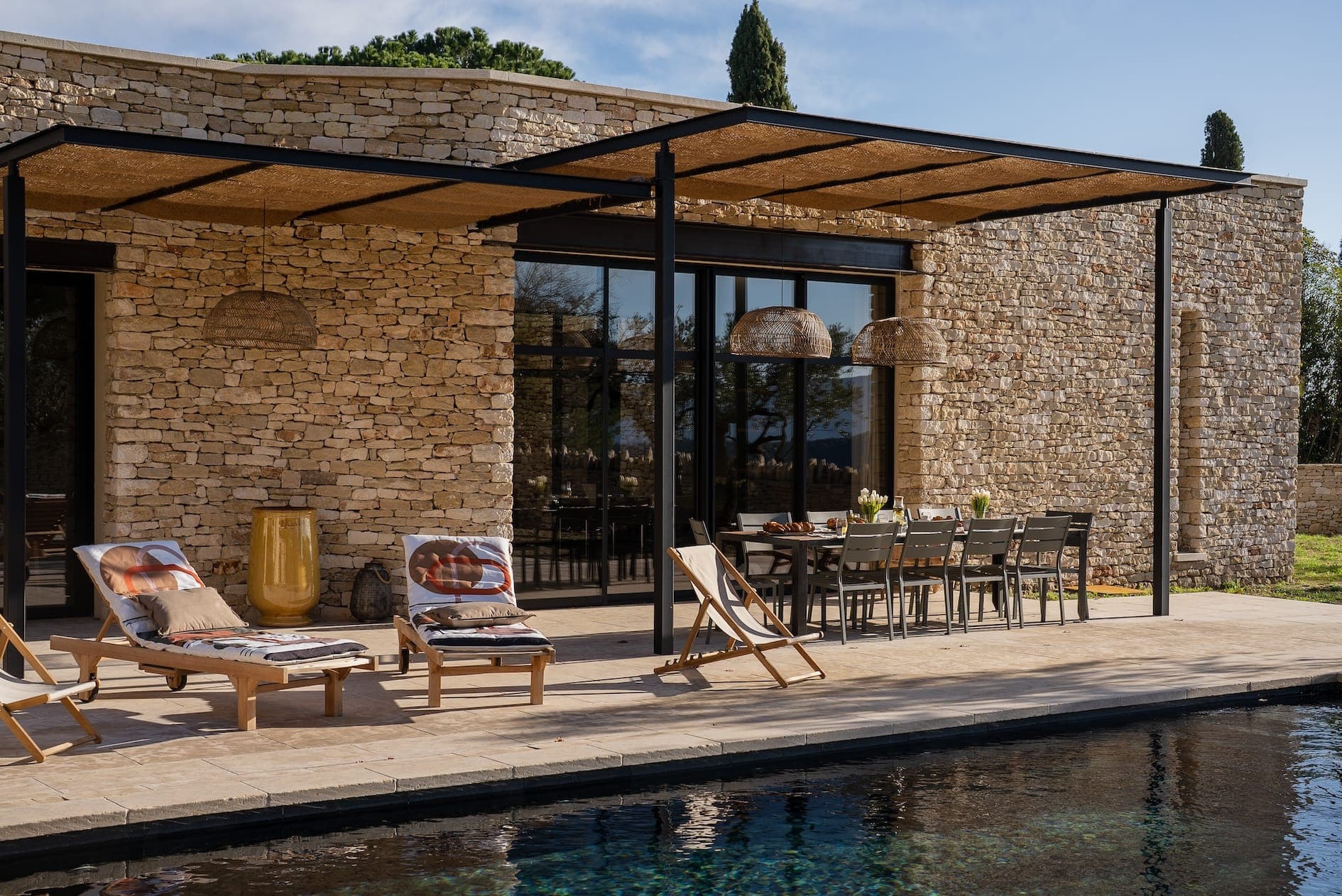 A stone villa with a modern outdoor patio featuring a long dining table and cushioned wooden lounge chairs by a pool. The area is shaded by a pergola, and the scene is set under a clear blue sky.